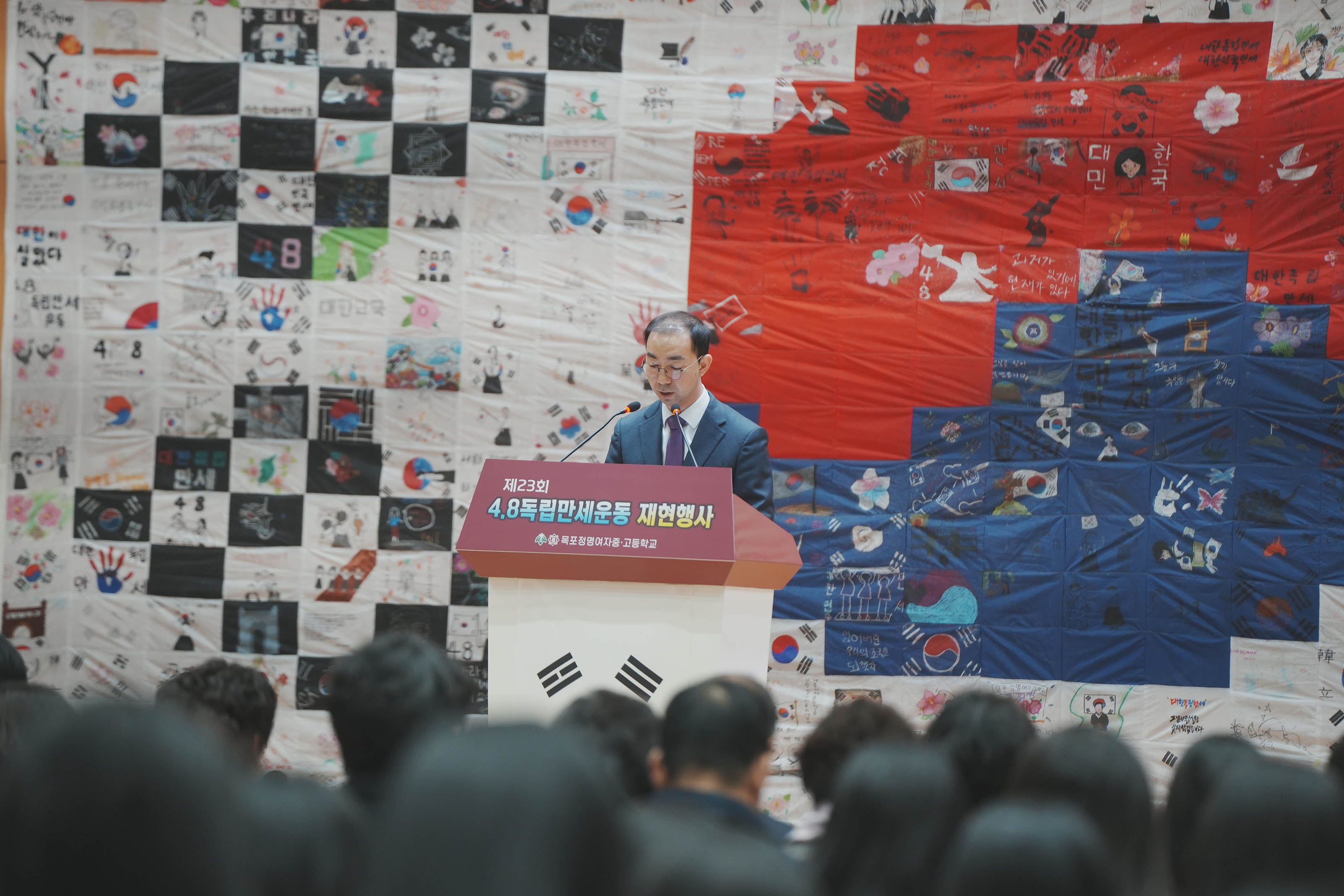 목포정명여자 중,고등학교에서 진행한 제23회 4.8독립만세운동 재현행사에서 보라색 넥타이를 맨 중년 남성이 단상앞에서 얘기하고 있다. 뒤로 태극기 작품이 보인다.
