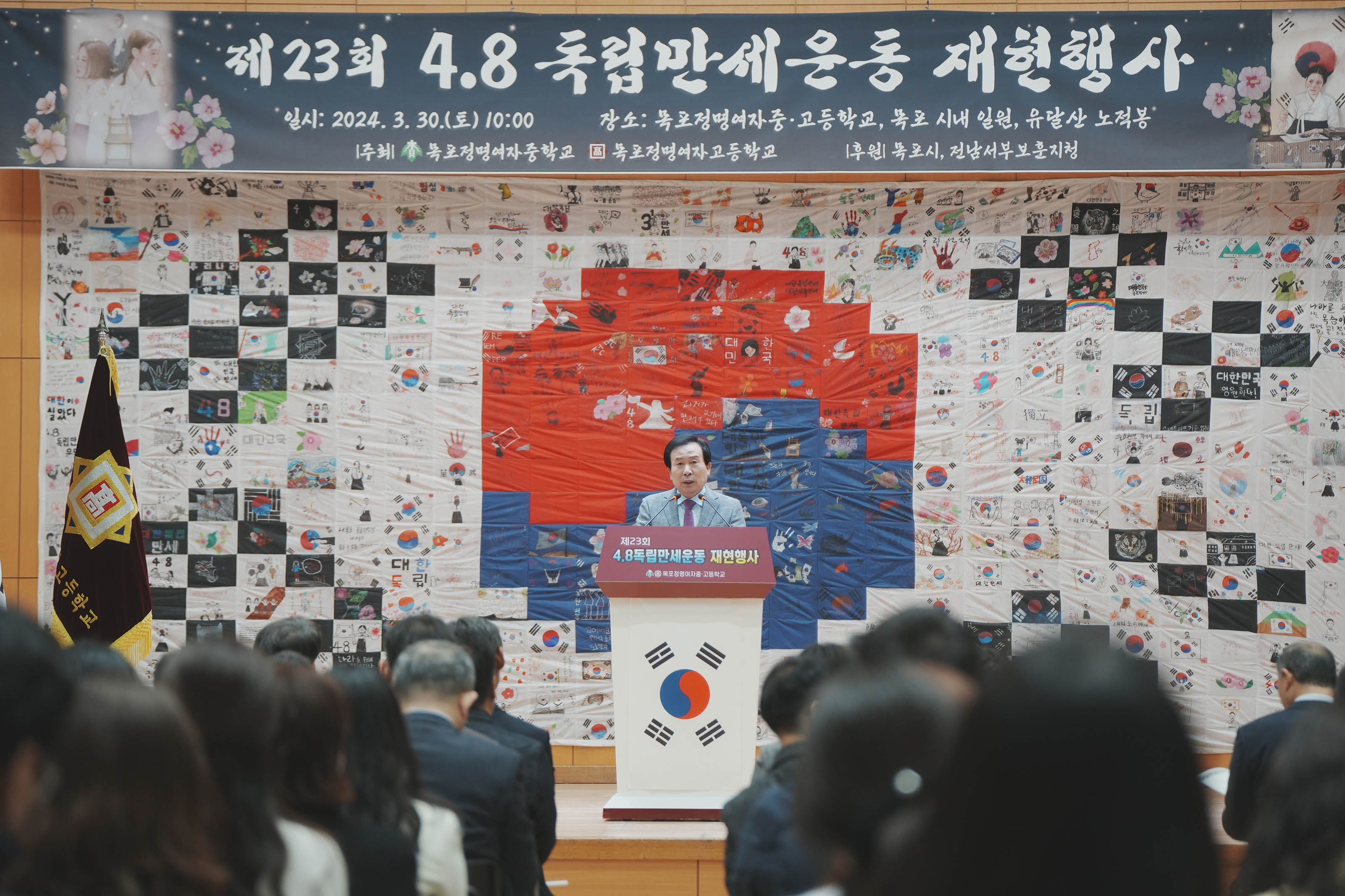 목포정명여자 중,고등학교에서 진행한 제23회 4.8독립만세운동 재현행사에서 박홍률 시장이 단상앞에서 얘기하고 있다. 뒤로 태극기 작품이 보인다. 박홍률 시장의 얘기를 듣고 있는 여러명의 사람들이 앉아있다.