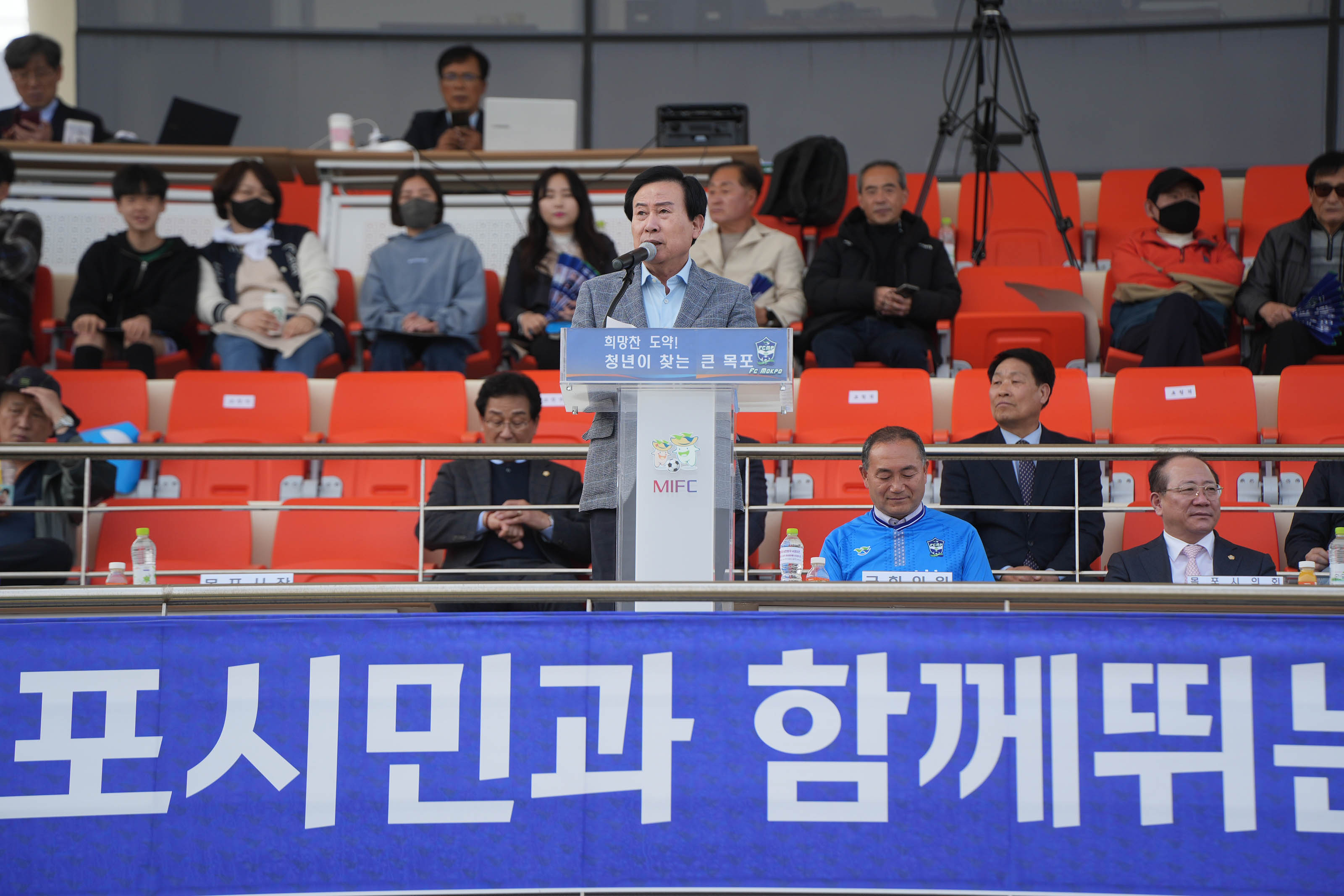 2024년 K3 홈 개막전에서 희망찬 도약! 청년이 찾는 큰 목포라고 적힌 단상에서 박홍률 시장이 마이크 앞에서 얘기를 하고 있다. 뒤로는 주황색 관중석 의자에 안자있는 사람들이 있다.