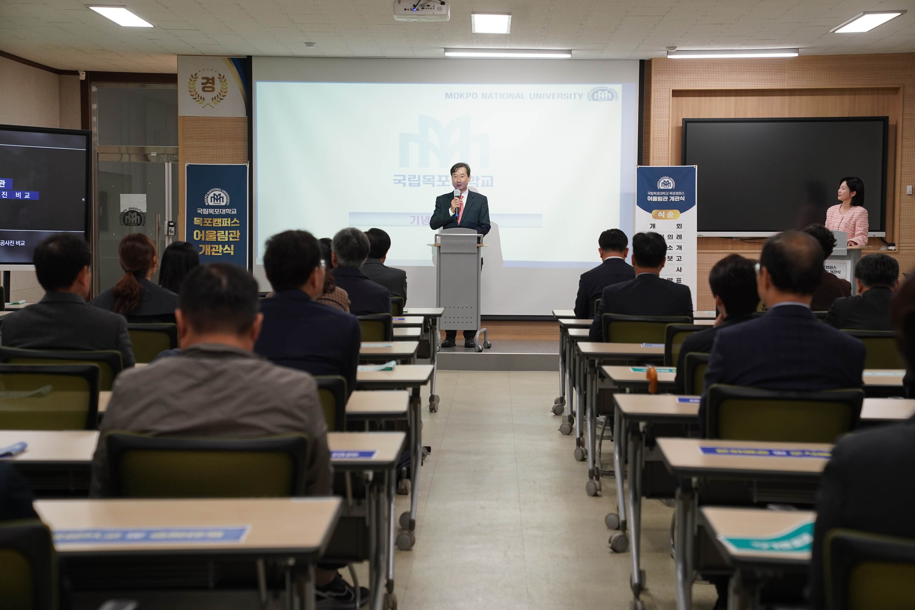 목포캠퍼스 어울림관 개관식에서 빨간 넥타이를 맨 남성이 마이크를 들고 단상에 서서 얘기하고 있고, 여러명의 남성들이 책상에 앉아 이야기를 듣고 있다.