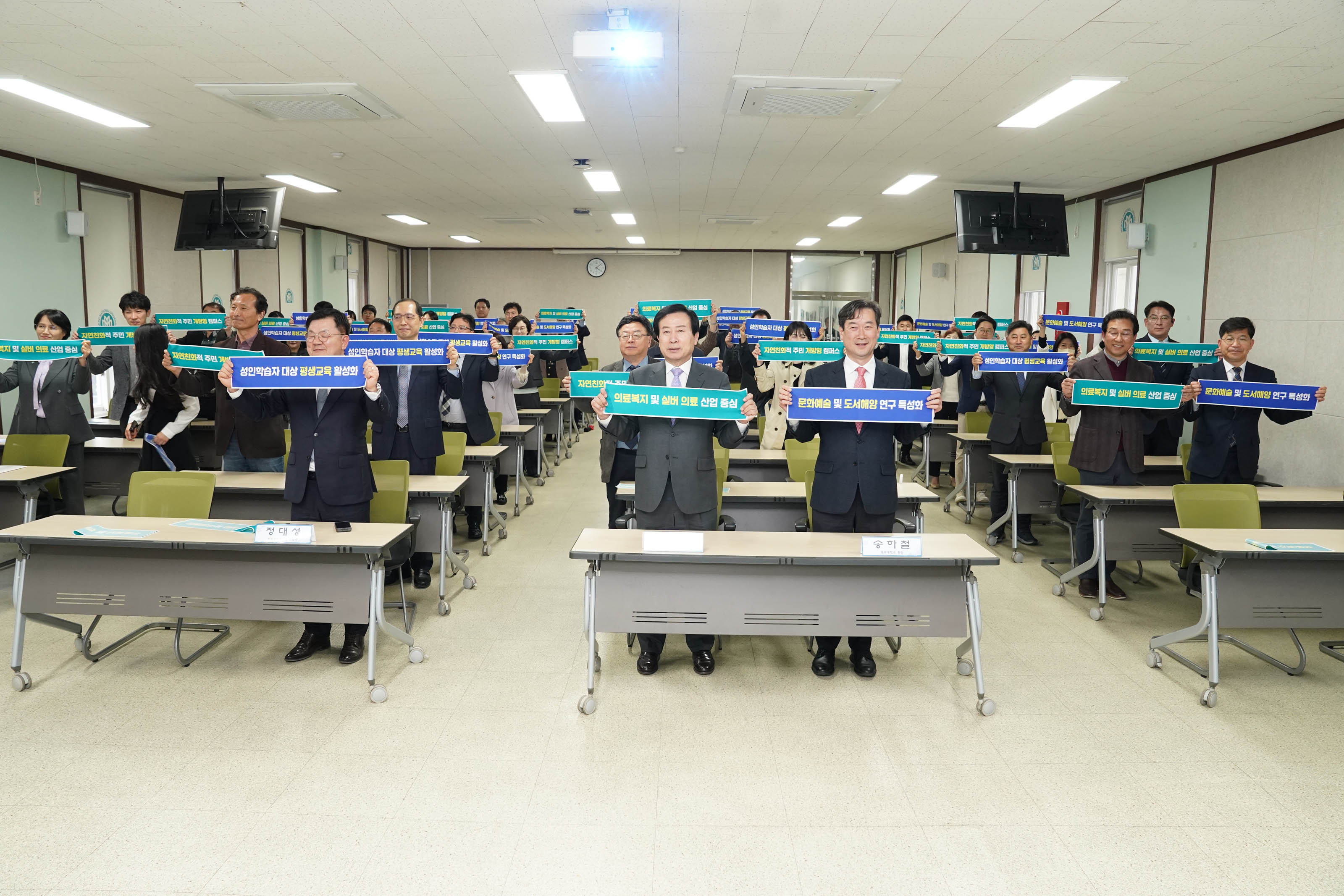 의료복지 및 실버 의료 산업 중심 플랜카드와 문화예술 및 도서해양 연구 특성화 플랜카드를 든 대략 40명 정도의 중년 남성과 중년 여성, 젊은 여성이 웃고있다.
