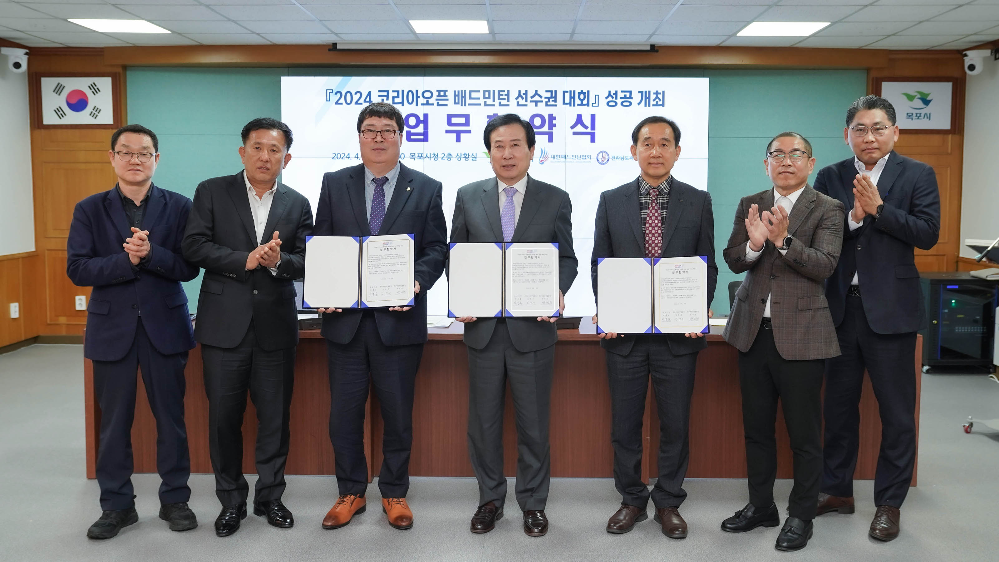 2024 코리아오픈 배드민턴 선수권 대회 성공 개최 업무협약식에서 김택규, 박홍률, 박계옥이 업무협약서를 들고 있고 양쪽에서 두명의 남성 총 네명이 박수 치고 있다.