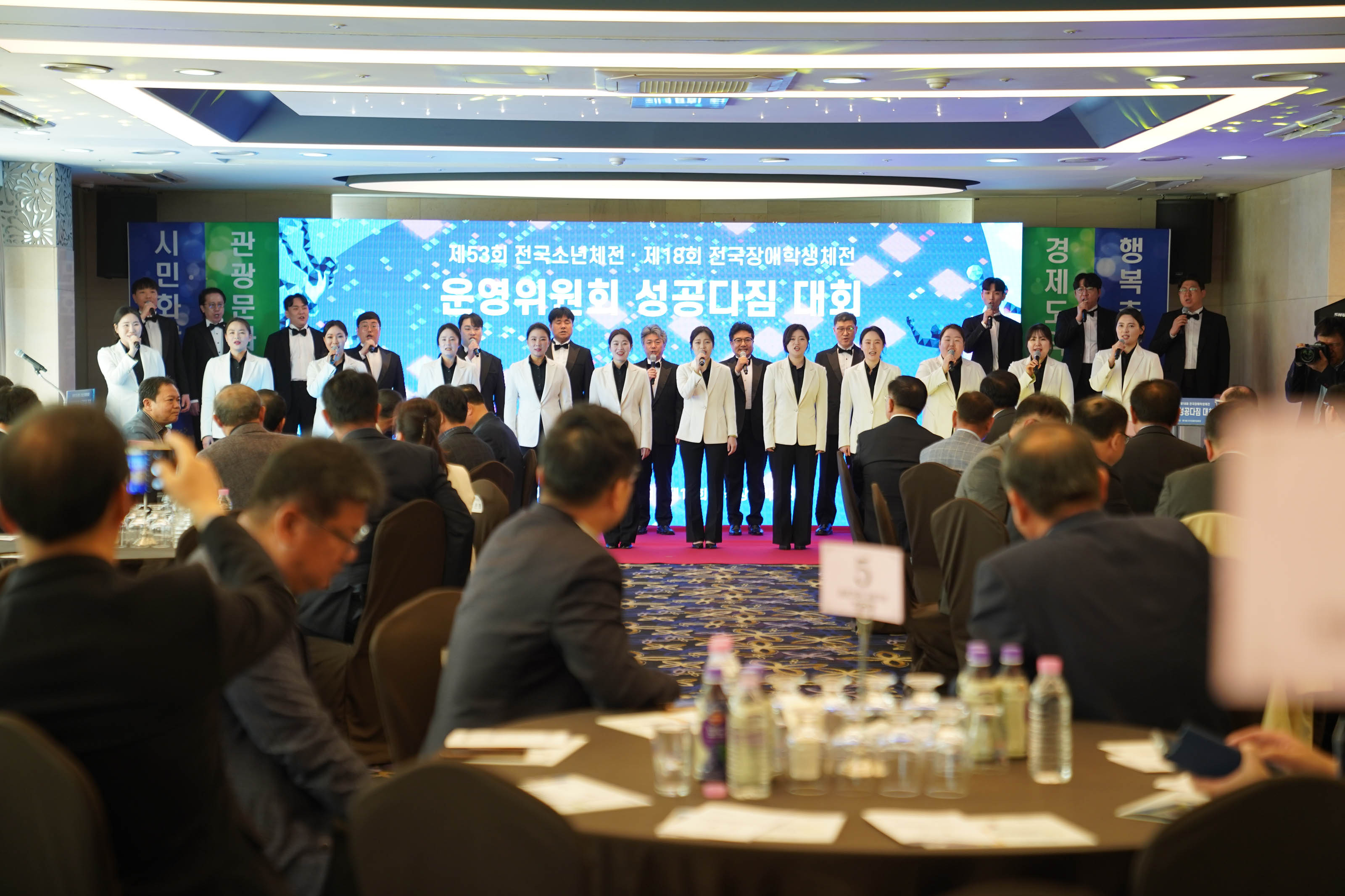 제53회 전국소년체전 제18회 전국장애학생체전 운영위원회 성공다짐 대회 글귀를 배경으로 노래를 부르며 무대를꾸미고있는 사람들과 앉아서 보고있는 참석자들