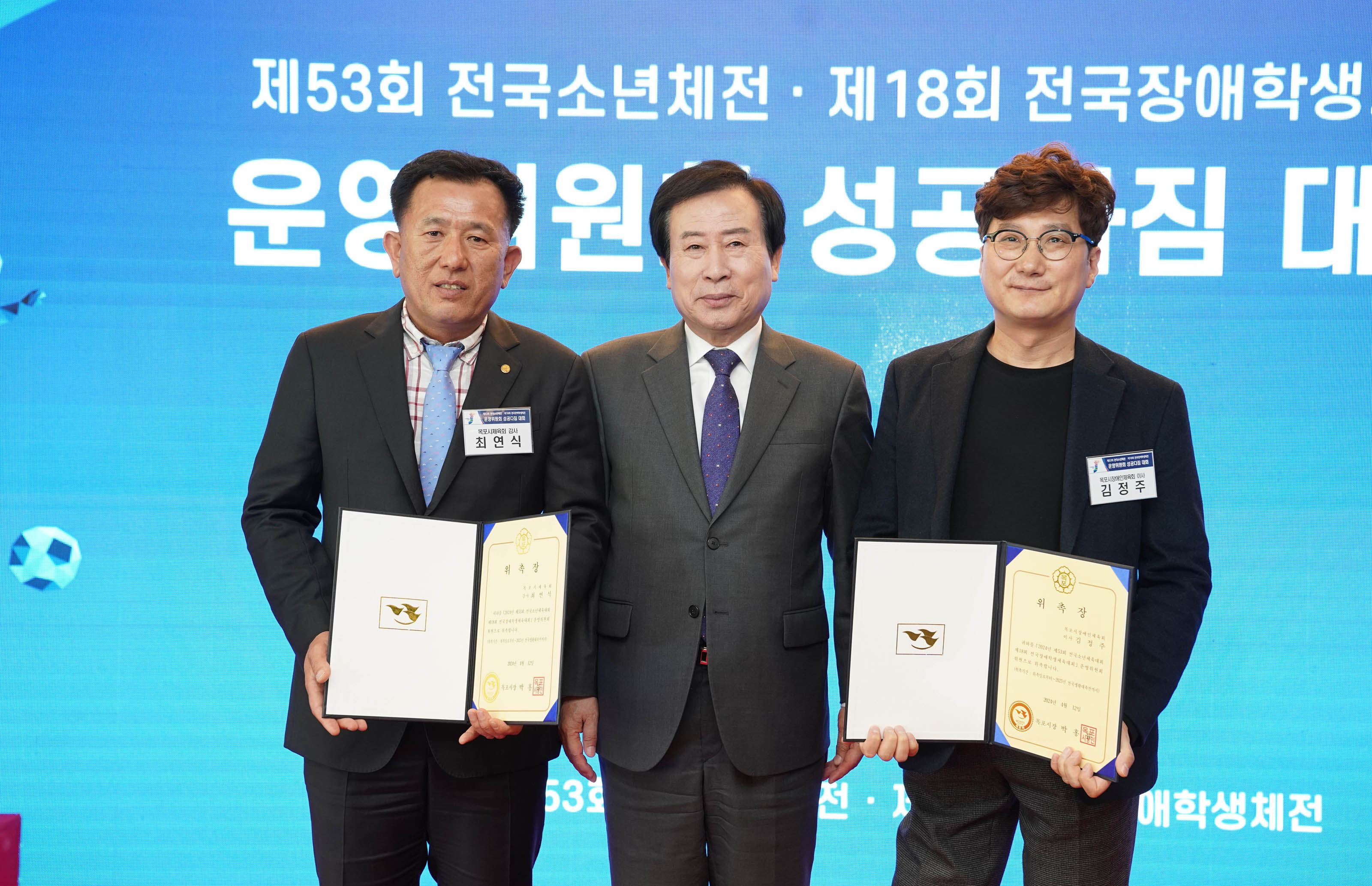 제53회 전국소년체전 제18회 전국장애학생체전 운영위원회 성공다짐 대회 글귀를 배경으로 수여받은 위축장을 들고 박홍률 시장 양쪽에서서 사진찍는 최연식, 김정주 이름표를 왼쪽 가슴에 달고있다.