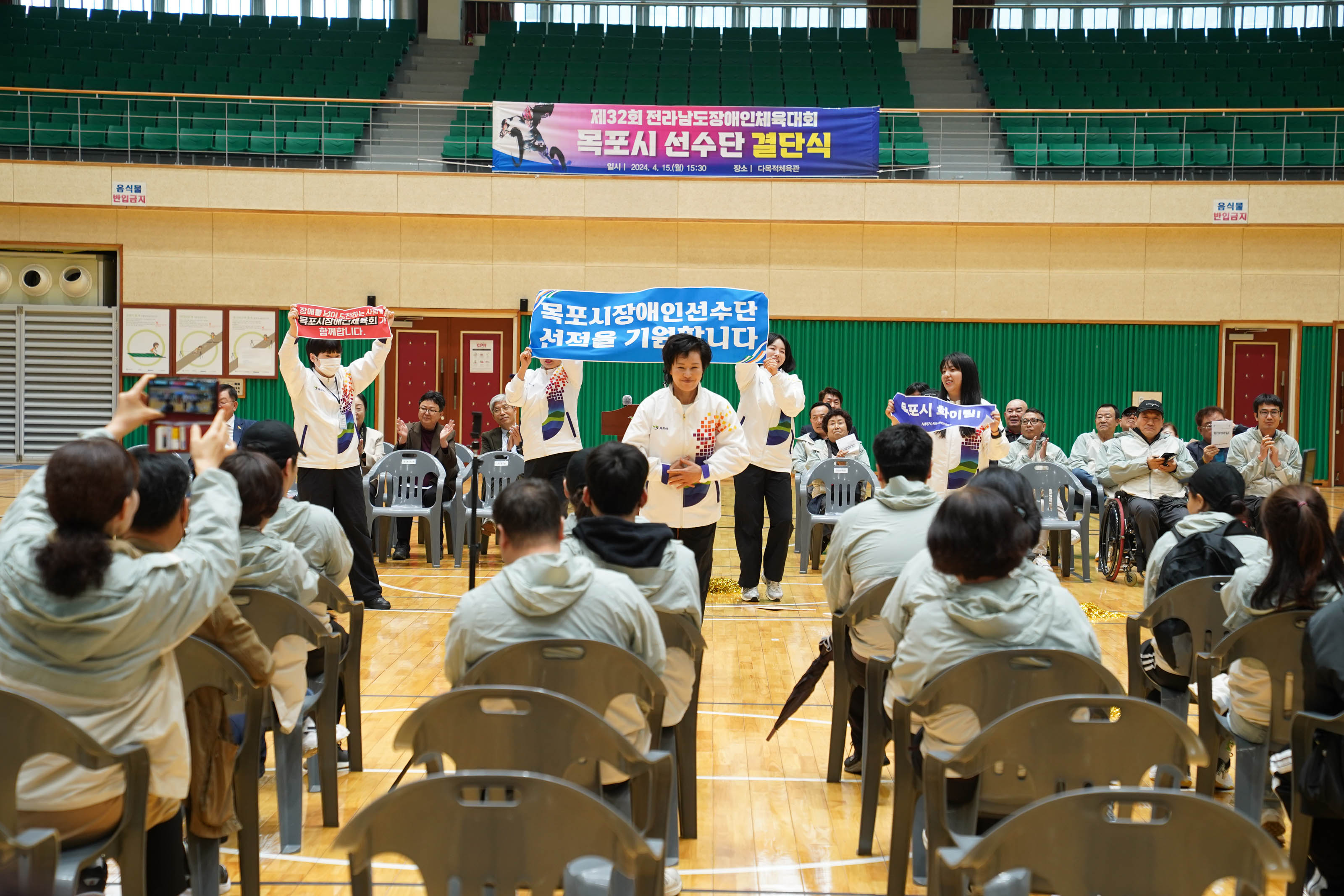 제32회 전라남도장애인체육대회 목포시 선수단 결단식 현수막이 체육관 뒤쪽편에 걸려있고 앞에서 목포시장애인선수단선점을 기원합니다 플랜카드를 들고 있고 그앞에 인사하고있는 관계자와 참석자들 뒷모습