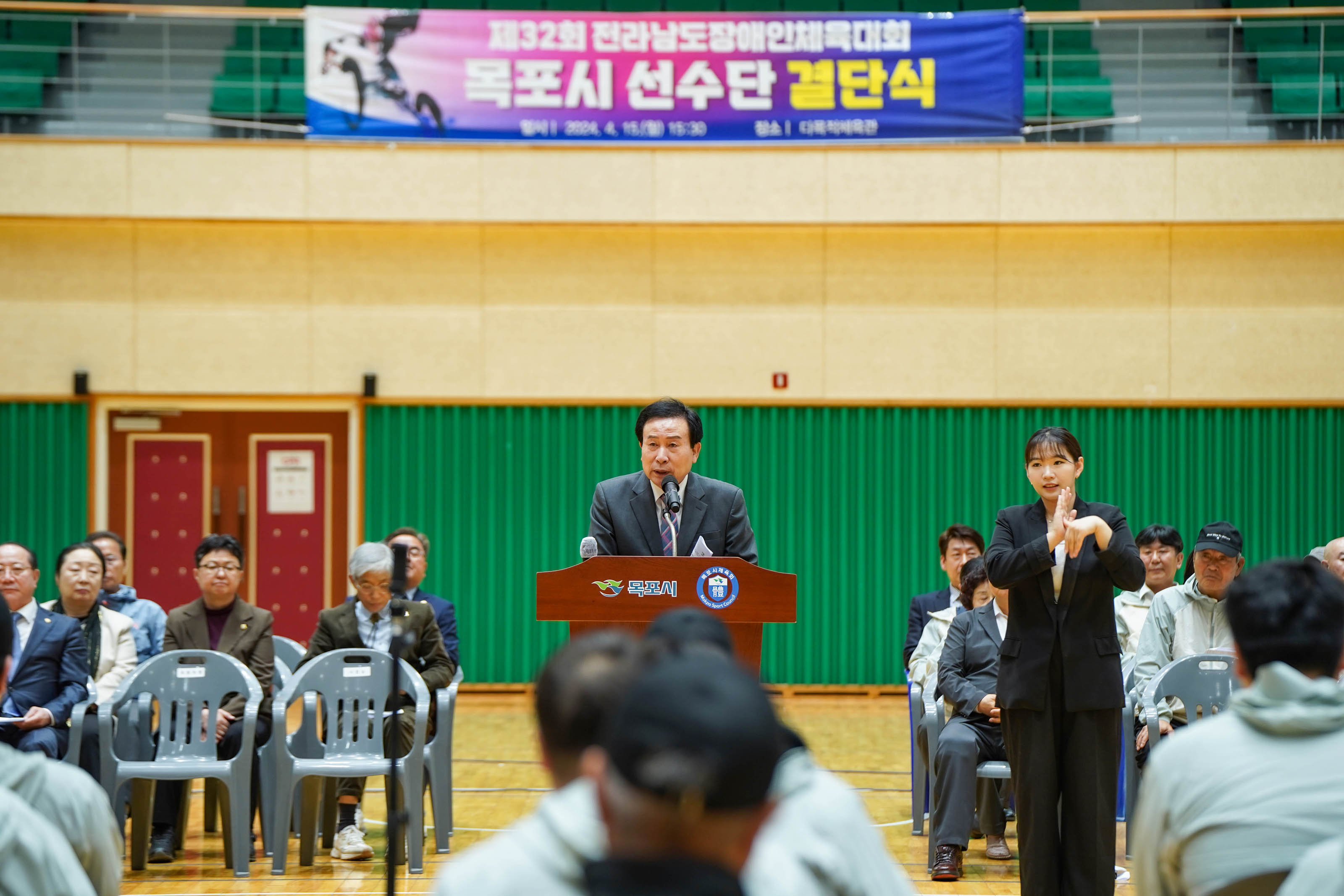 제32회 전라남도장애인체육대회 목포시 선수단 결단식 현수막이 체육관 뒤쪽편에 걸려있고 목포시가 적현있는 단상에서 연설하고있는 박홍률 시장과 옆에서 수화통역을 하고있고 경청하고 있는 참석자들