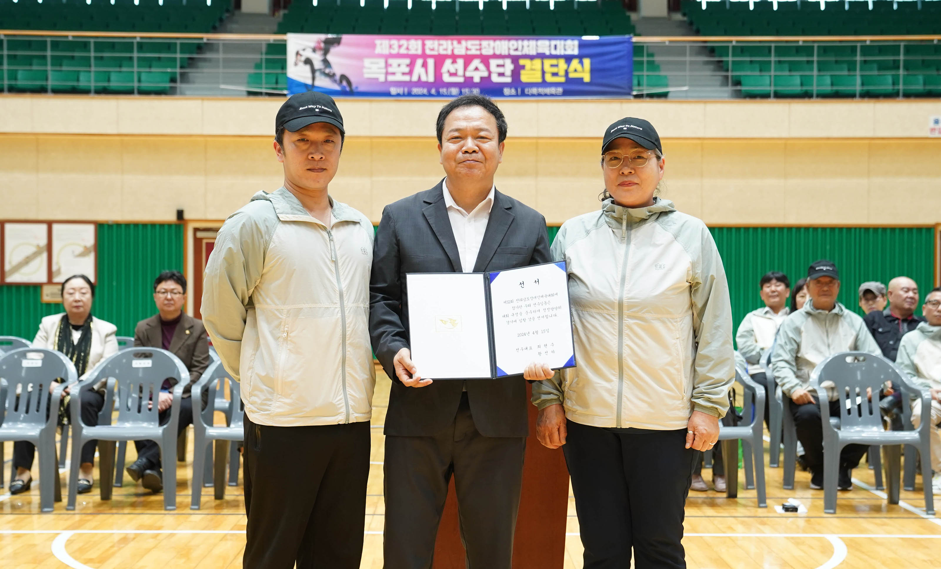 제32회 전라남도장애인체육대회 목포시 선수단 결단식 현수막이 체육관 뒤쪽편에 걸려있고 선서장을 들고 있는 관계자와 양옆에서있는 다른 관계자들 뒤로 참석자들이 앉아있다