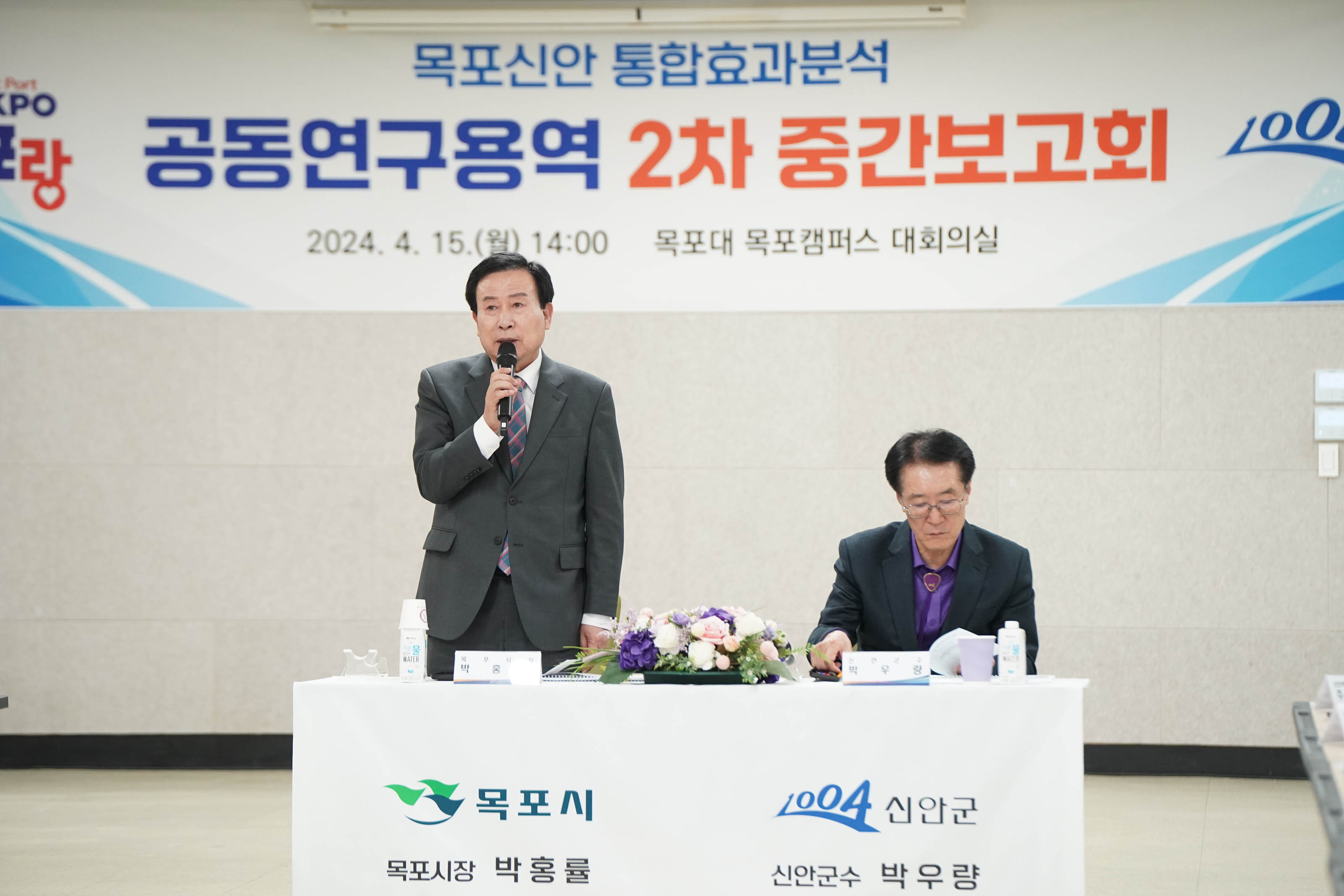 목포신안 통합효과분석 공동연구용역 2차 중간보고회 2024. 4.15(월) 14:00 목포대 목포캠퍼스 대회의실 현수막을 배경으로 목포시 목포시장 박홍률 신안군 신안군수 박우량이 적혀있는 단상에 박우량 군수는 앉아있고 박홍률 시장은 일어서서 마이크를 들고 발표를 하고있다.