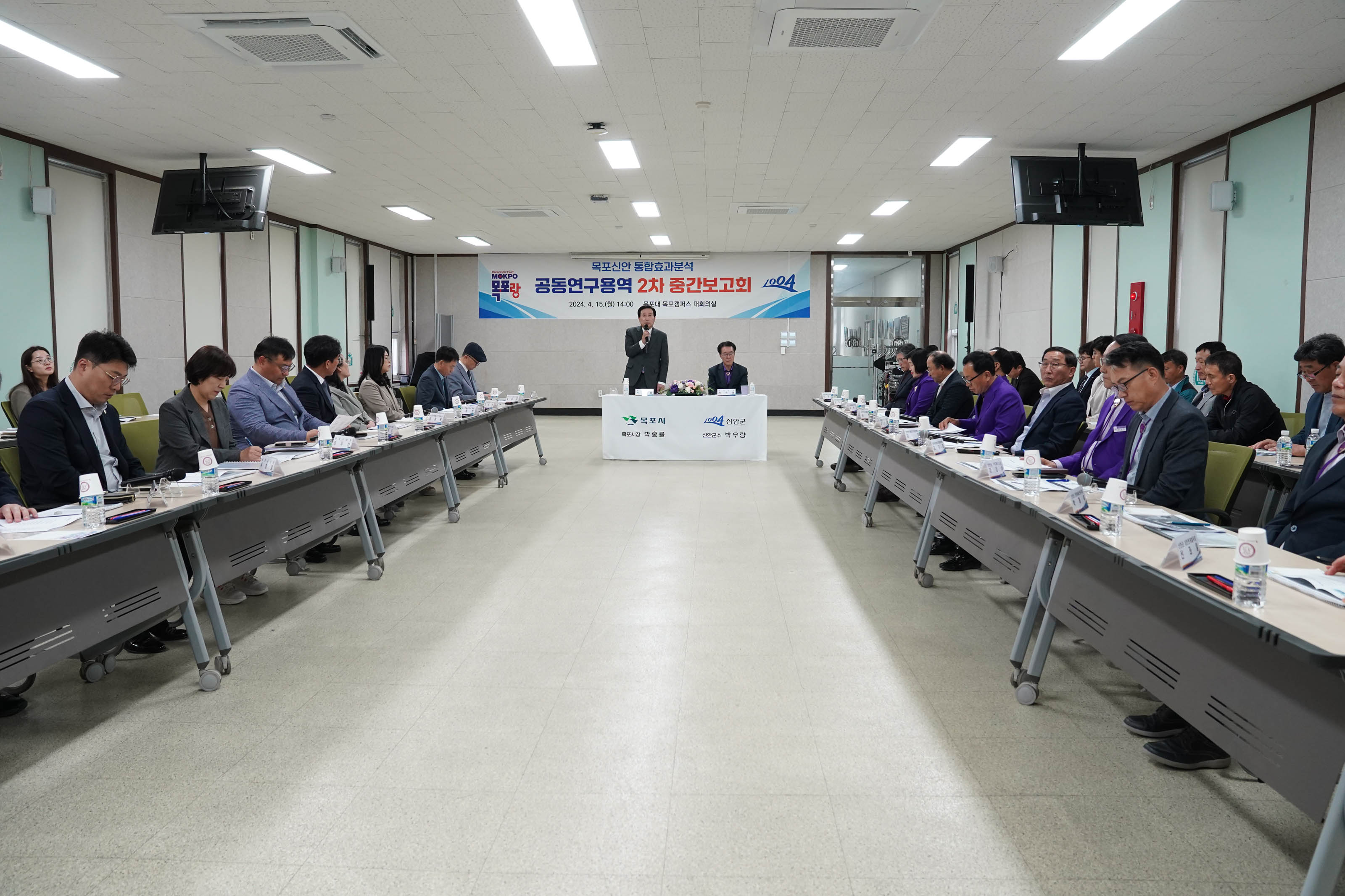 목포신안 통합효과분석 공동연구용역 2차 중간보고회 2024. 4.15(월) 14:00 목포대 목포캠퍼스 대회의실 현수막을 배경으로 목포시 목포시장 박홍률 신안군 신안군수 박우량이 적혀있는 단상에 박우량 군수는 앉아있고 박홍률 시장은 일어서서 마이크를 들고 발표를 하고있고 경청하는 관계자들 멀리서 찍은사진