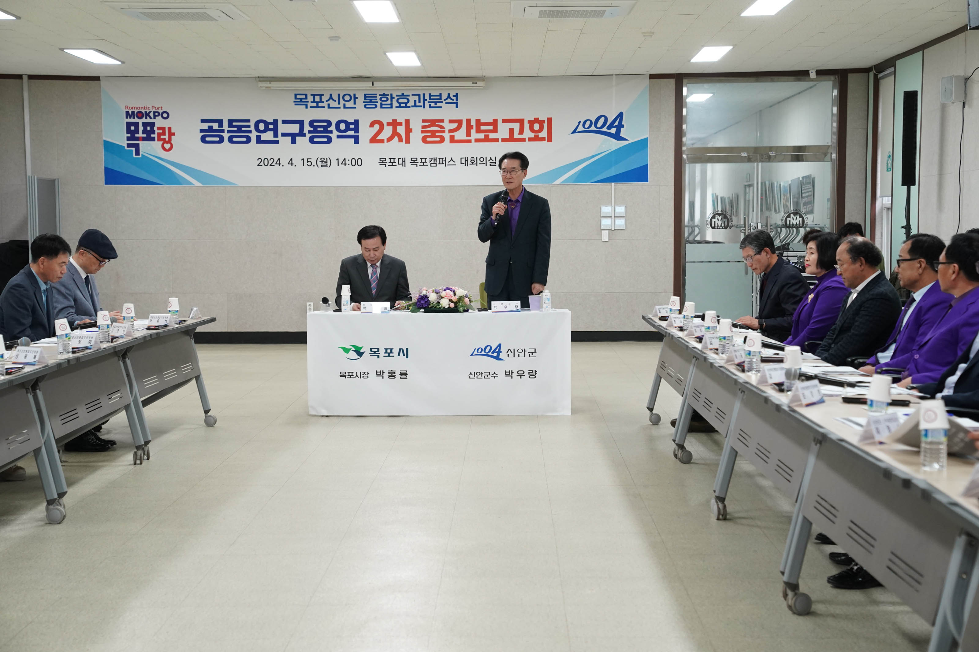 목포신안 통합효과분석 공동연구용역 2차 중간보고회 2024. 4.15(월) 14:00 목포대 목포캠퍼스 대회의실 현수막을 배경으로 목포시 목포시장 박홍률 신안군 신안군수 박우량이 적혀있는 단상에 박홍률 시장은 앉아있고 박우량 군수는 일어서서 마이크를 들고 발표를 하고있다.