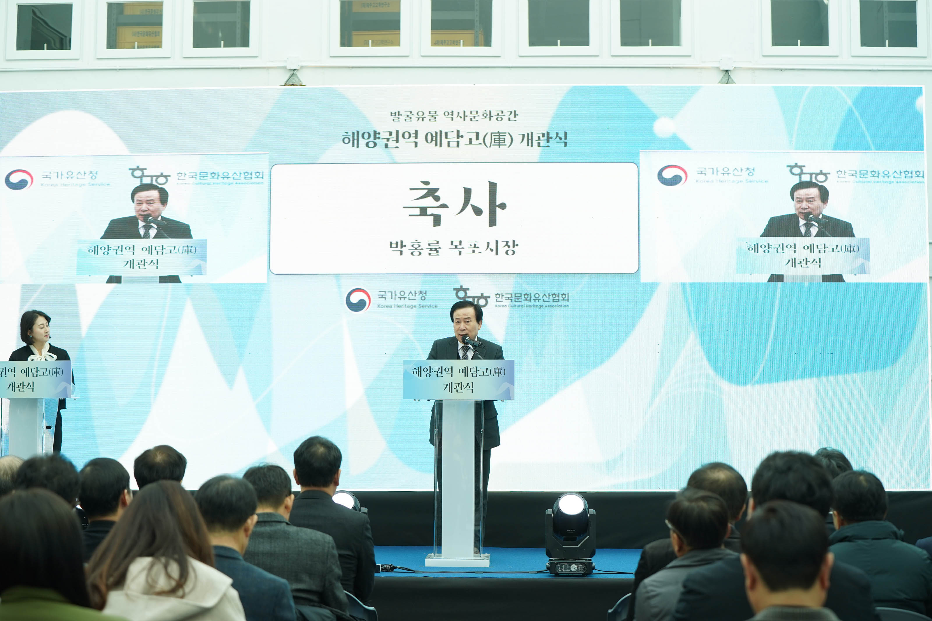 배경에 ‘발굴유물 역사문화공간 해양권역 예담고(庫) 개관식’ 이라는 문구가 적혀 있으며, 단상 중앙에서 박홍률 목포시장이 축사를 하고 있는 모습. 양옆 스크린에는 축사 장면이 실시간으로 송출되고 있고, 앞쪽에는 여러 명의 참석자가 앉아 있는 모습.
