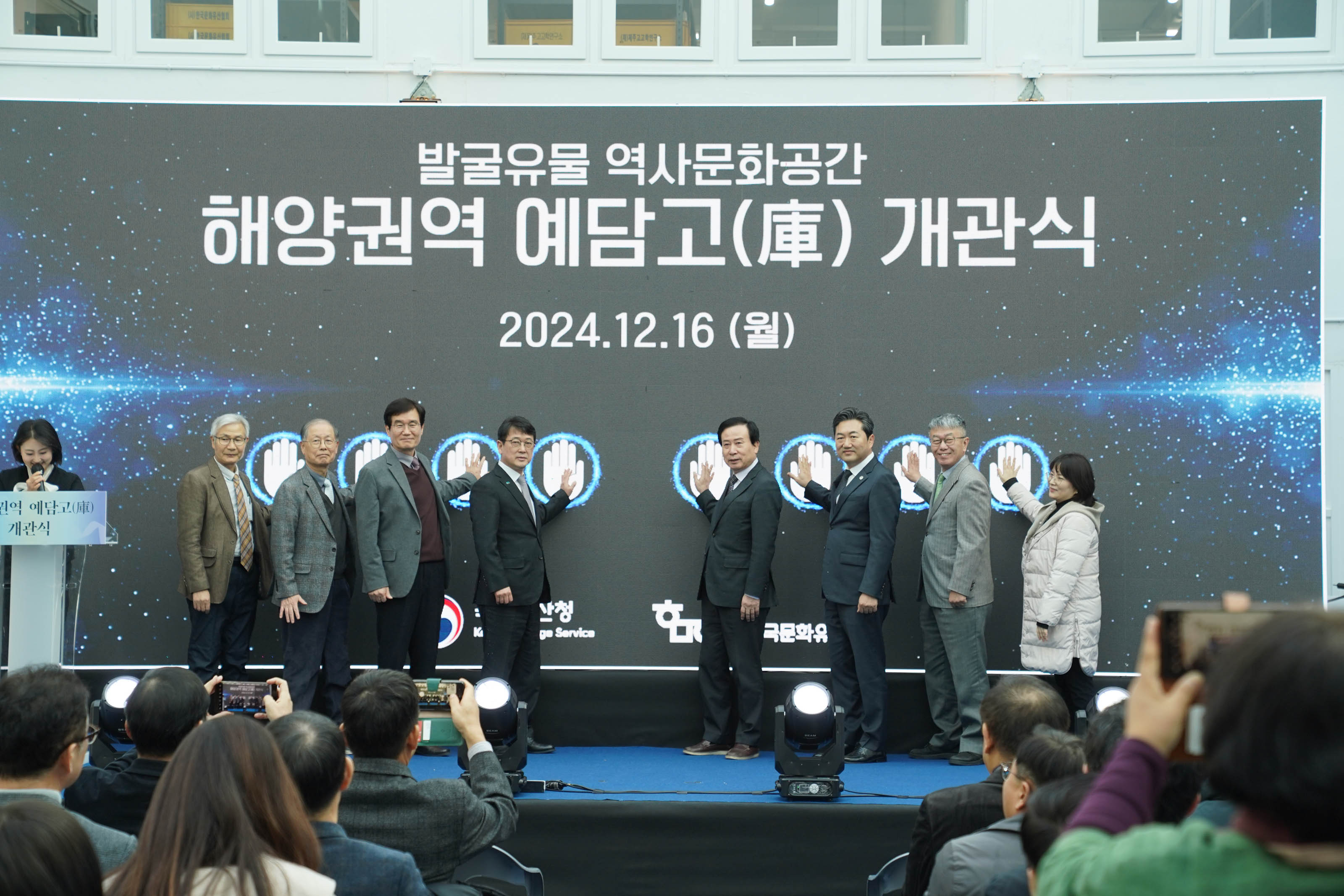 ‘발굴유물 역사문화공간 해양권역 예담고(庫) 개관식 2024.12.16 (월)’이라고 적힌 대형 전광판 앞에서 8명의 관계자들이 손바닥 모양의 터치패드에 손을 올리며 개관 퍼포먼스를 진행하고 있는 모습. 무대 아래에는 관람객들이 해당 장면을 촬영하고 있음.