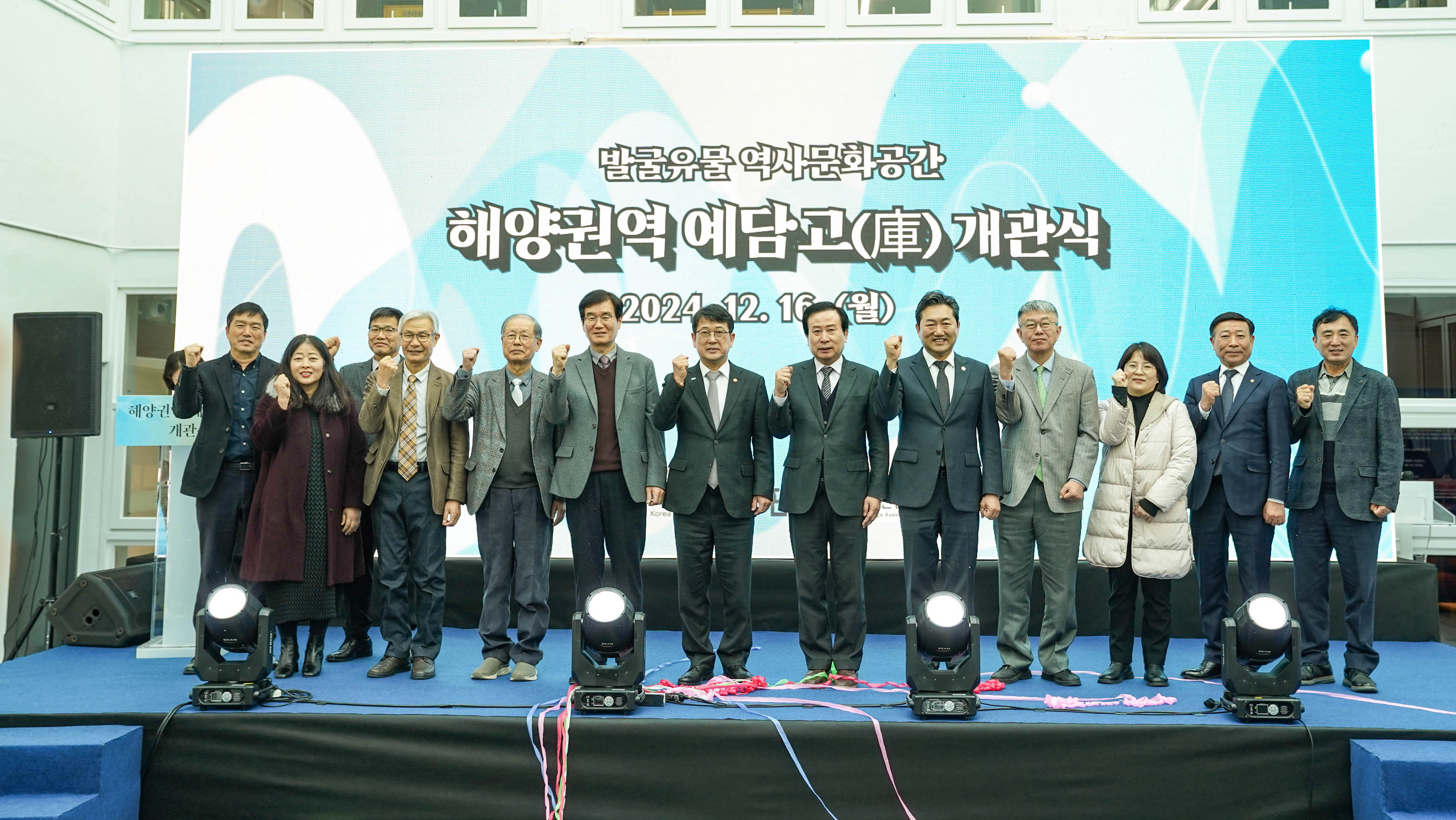 ‘발굴유물 역사문화공간 해양권역 예담고(庫) 개관식2024.12.16(월)' 이라는 문구가 적힌 배경 앞 단상에서 13명의 관계자들이 줄지어 서서 주먹을 쥐고 포즈를 취하며 단체 기념사진을 찍고 있는 모습.