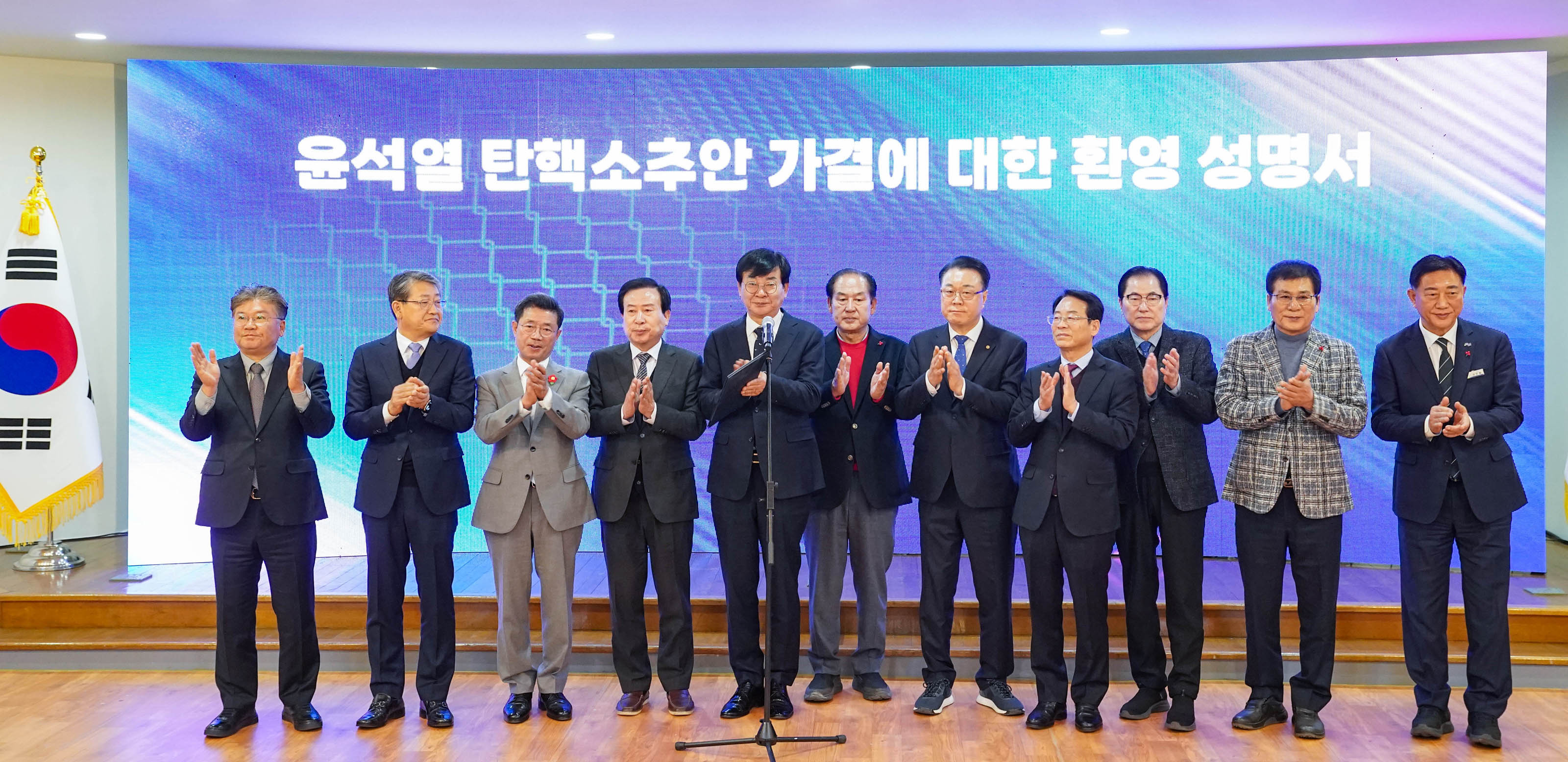 '윤석열 탄핵소추안 가결에 대한 환영 성명서'라는 문구가 적힌 대형 전광판 앞에서 정장 차림의 인사 11명이 일렬로 서서 박수를 치고 있는 모습. 무대 왼쪽에는 태극기가 세워져 있다.