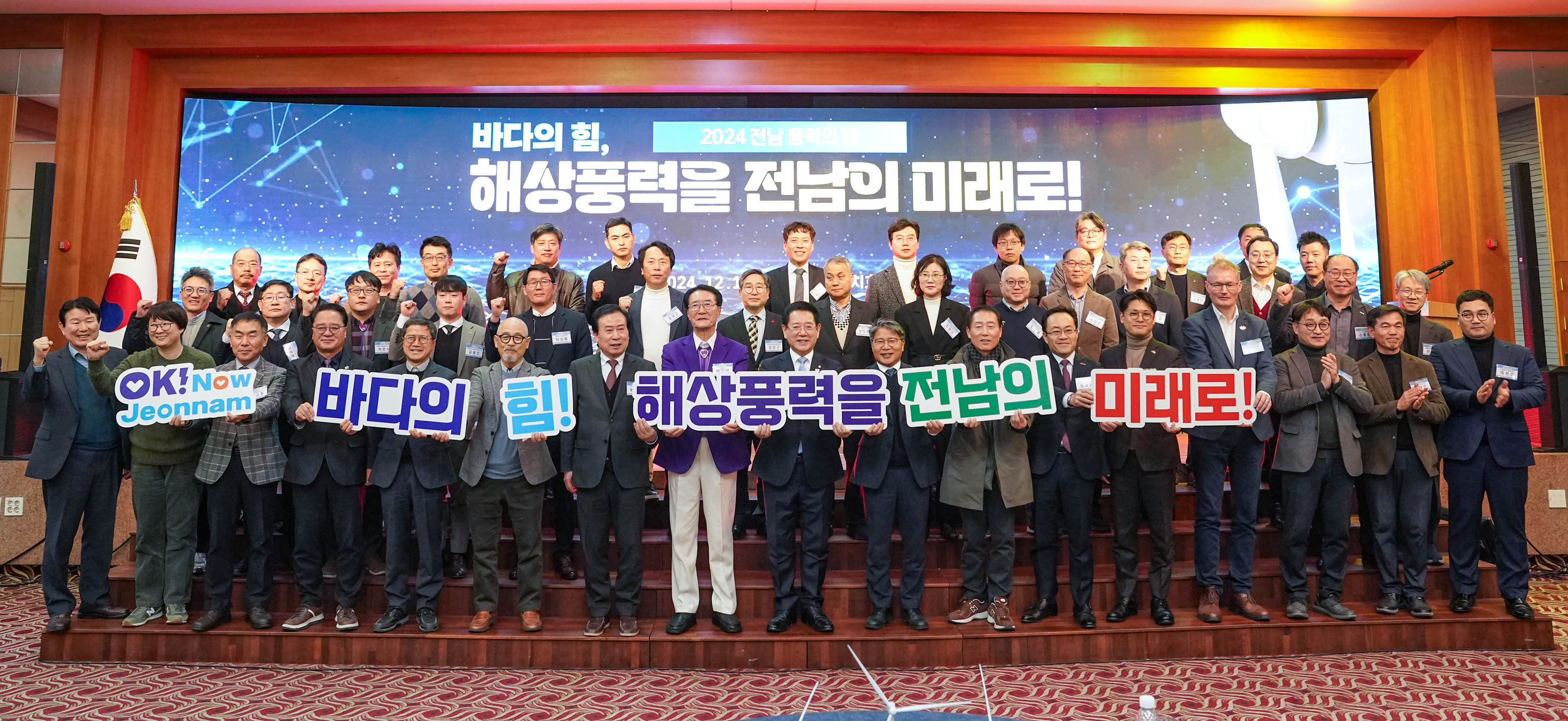 '2024 전남 풍력의 밤' 행사에서 '바다의 힘, 해상풍력을 전남의 미래로!'라는 문구가 적힌 대형 전광판 앞에 약 50여 명의 참가자들이 단체 기념사진을 촬영하고 있는 모습. 앞줄 인사들은 'OK! Now Jeonnam', '바다의 힘!', '해상풍력을', '전남의', '미래로!' 등의 문구가 적힌 피켓을 들고 있다.