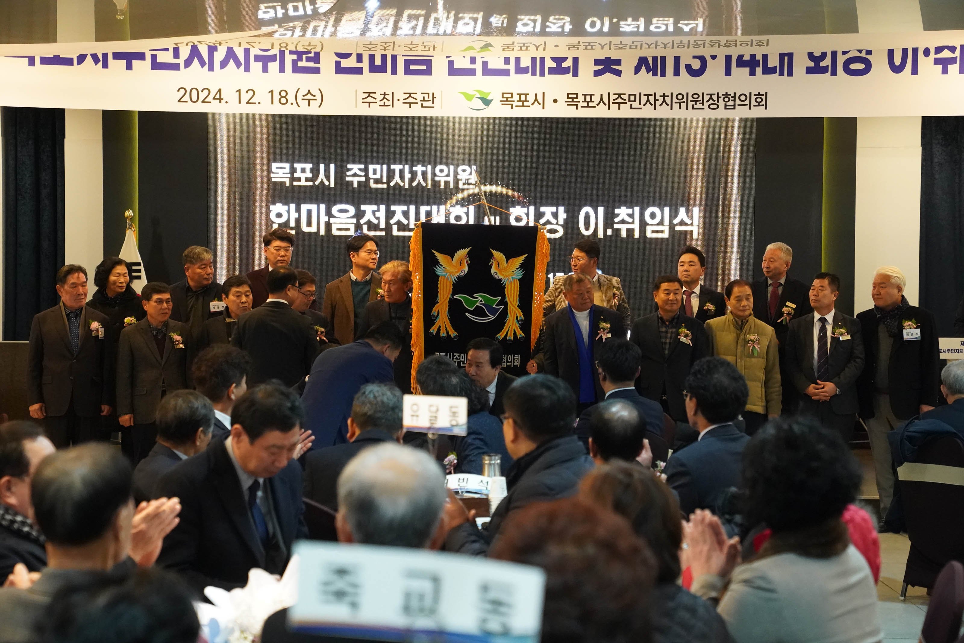 '2024년 12월 18일(수) |주최 · 주관| 목포시 · 목포시주민자치위원장협의뢰' 라는 현수막이 걸려있고 대형 전광판에는 '목포시 주민자치위원회 한마음전진대회 회장 이,취임식'이라는 문구가 적혀있다. 무대 위에는 깃발을 중심으로 여러 명의 위원들이 단상에 나란히 서 있다. 무대 아래에는 수십 명의 참석자들이 앉아 행사를 지켜보고 있다.