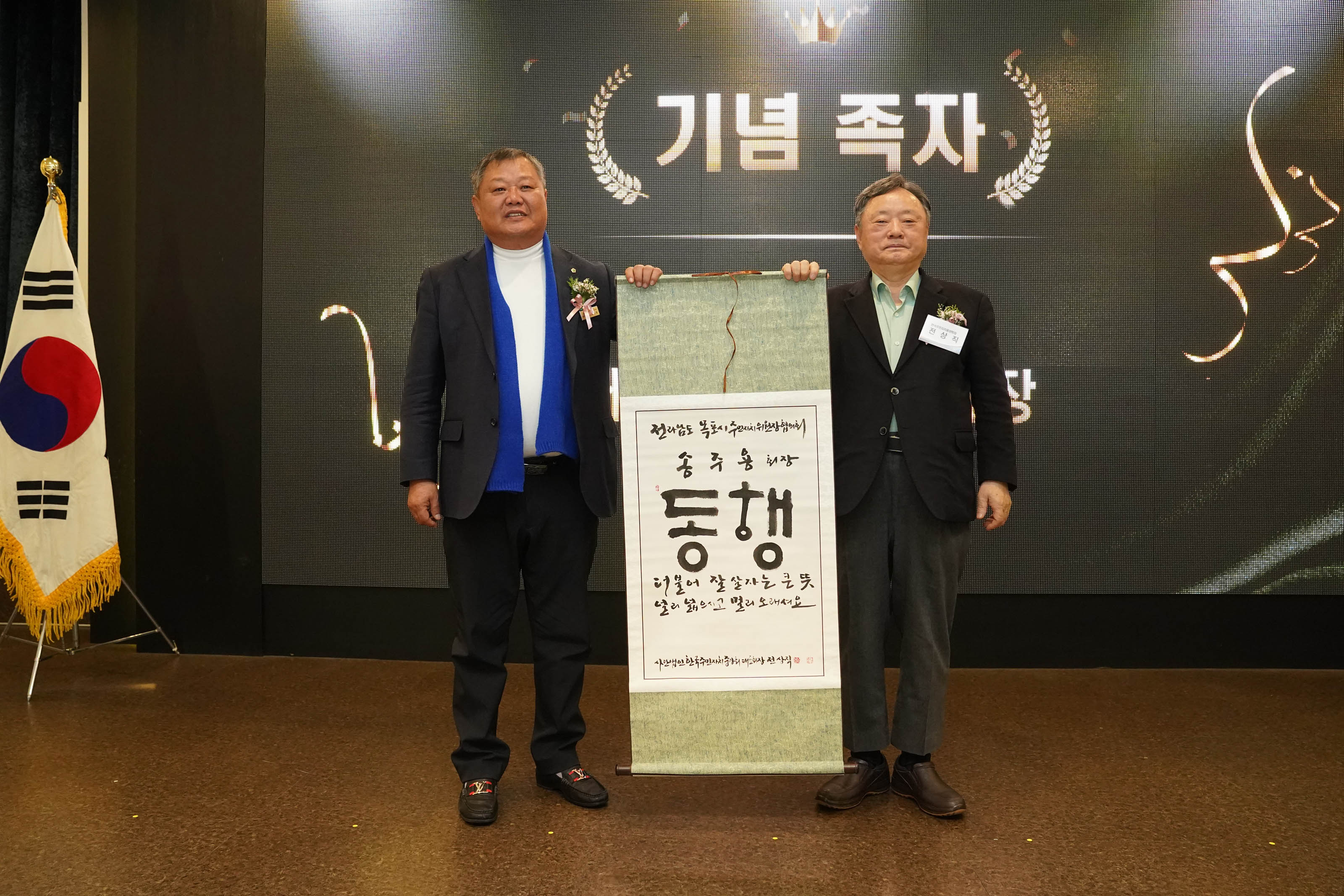 '기념 족자'라는 문구가 적힌 배경 앞에서 두 명의 관계자가 족자를 함께 들고 서 있는 모습. 족자에는 '동행'이라는 글씨와 함께 손글씨로 작성된 문구가 적혀 있으며, 왼쪽에는 태극기가 세워져 있다.