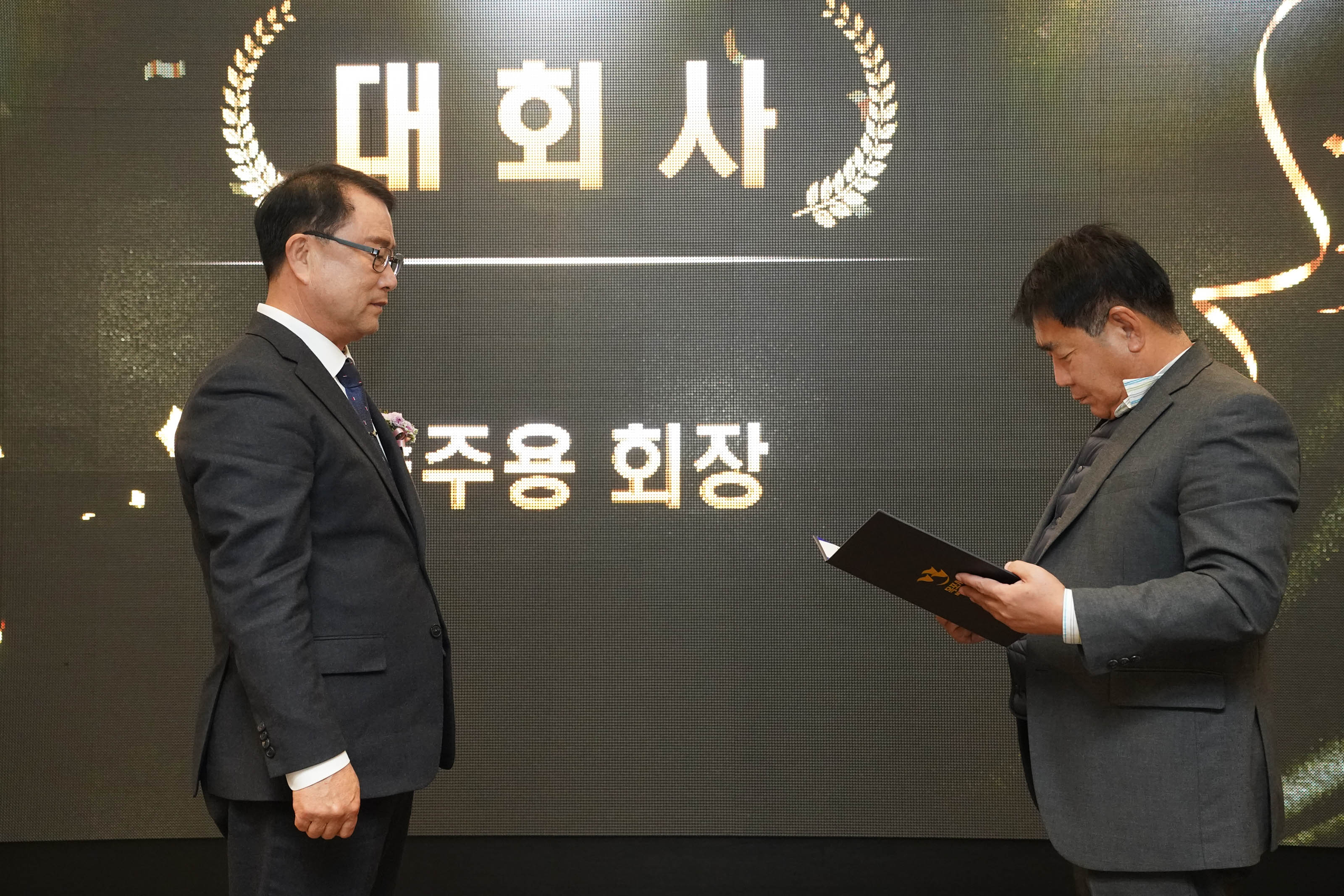 '대회사'라는 문구가 적힌 전광판 앞에서 신임 회장의 취임식이 진행되고 있는 모습. 오른쪽의 관계자가 취임사를 읽고 있고, 왼쪽의 남성은 단정한 정장 차림으로 마주 서 있다.