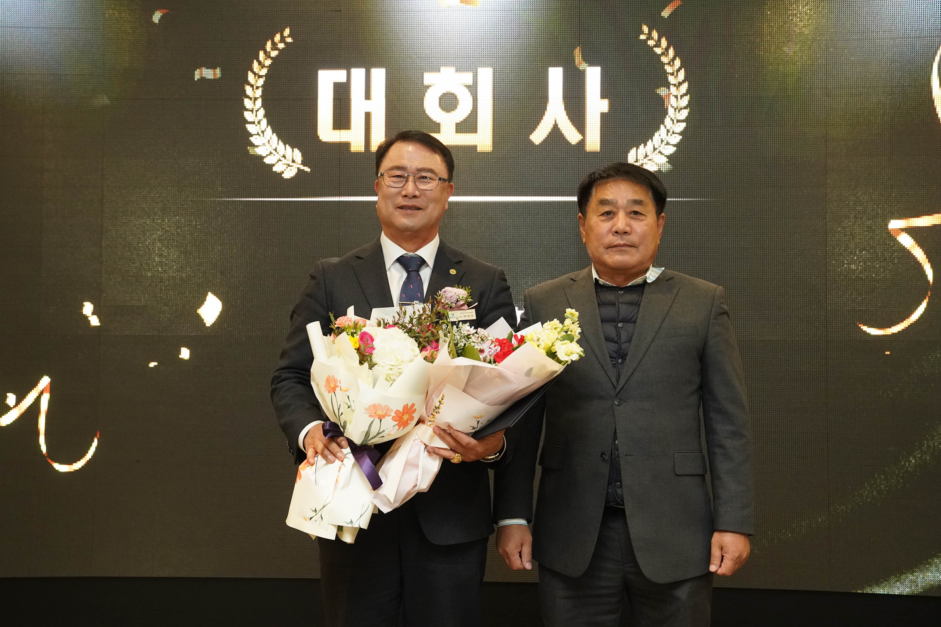 ‘대회사’라는 문구가 적힌 배경 앞에서 두 관계자가 나란히 서서 기념사진을 촬영하고 있는 모습. 신임 회장은 정장을 입고 꽃다발을 두 개 들고 있으며, 오른쪽 남성은 차분한 표정으로 서 있다.