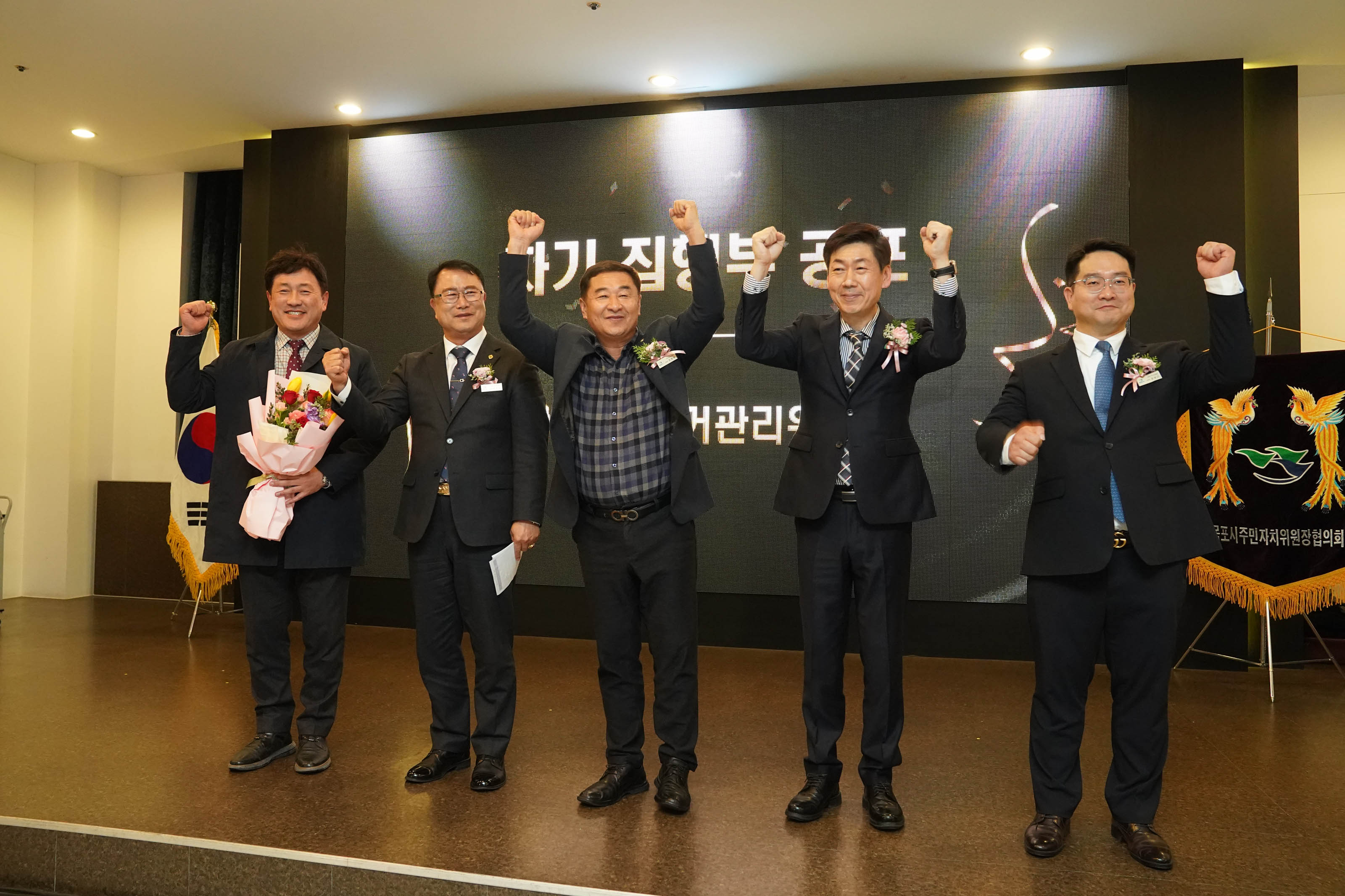 단상에서 다섯 명의 관계자들이 일제히 주먹을 쥐고 손을 들어올리며 파이팅을 외치고 있는 모습. 왼쪽 인물은 꽃다발을 들고 있으며, 모두 정장을 입고 가슴에 꽃을 달고 있어 공식적인 행사임을 나타낸다. 배경 양옆에는 태극기와 협회기를 비롯한 장식이 배치되어 있다.