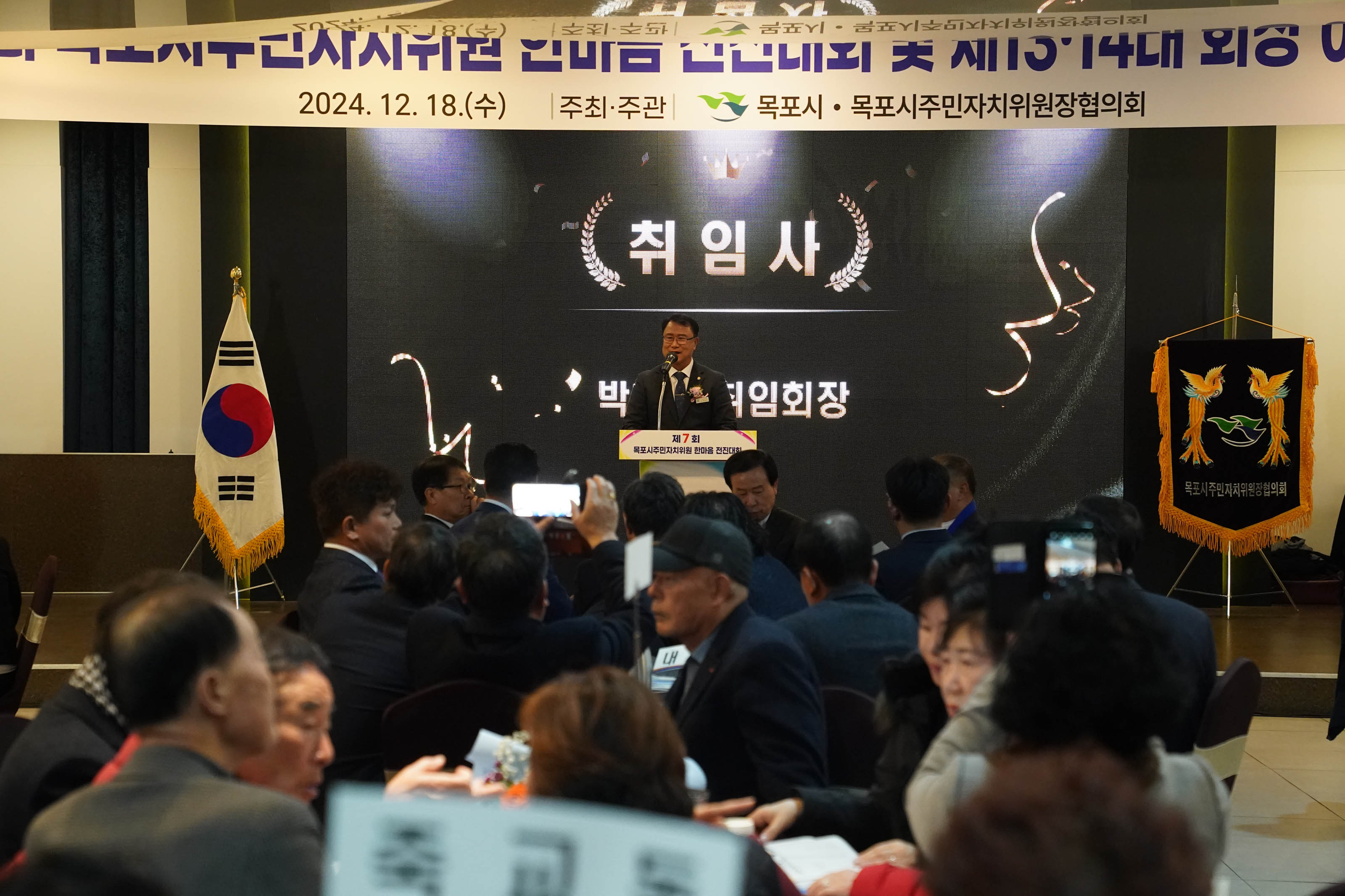'2024년 12월 18일 (수) |주최·주관| 목포시 주민자치위원장협의회' 라는 문구가 현수막에 적혀있고 ‘취임사’라는 문구가 적힌 대형 전광판 앞 단상에서 회장님이 마이크를 들고 취임사를 하고 있다. 단상 아래에는 취임식을 지켜보는 많은 참석자들이 앉아 있으며, 태극기와 협회 깃발이 양옆에 배치되어 있다.