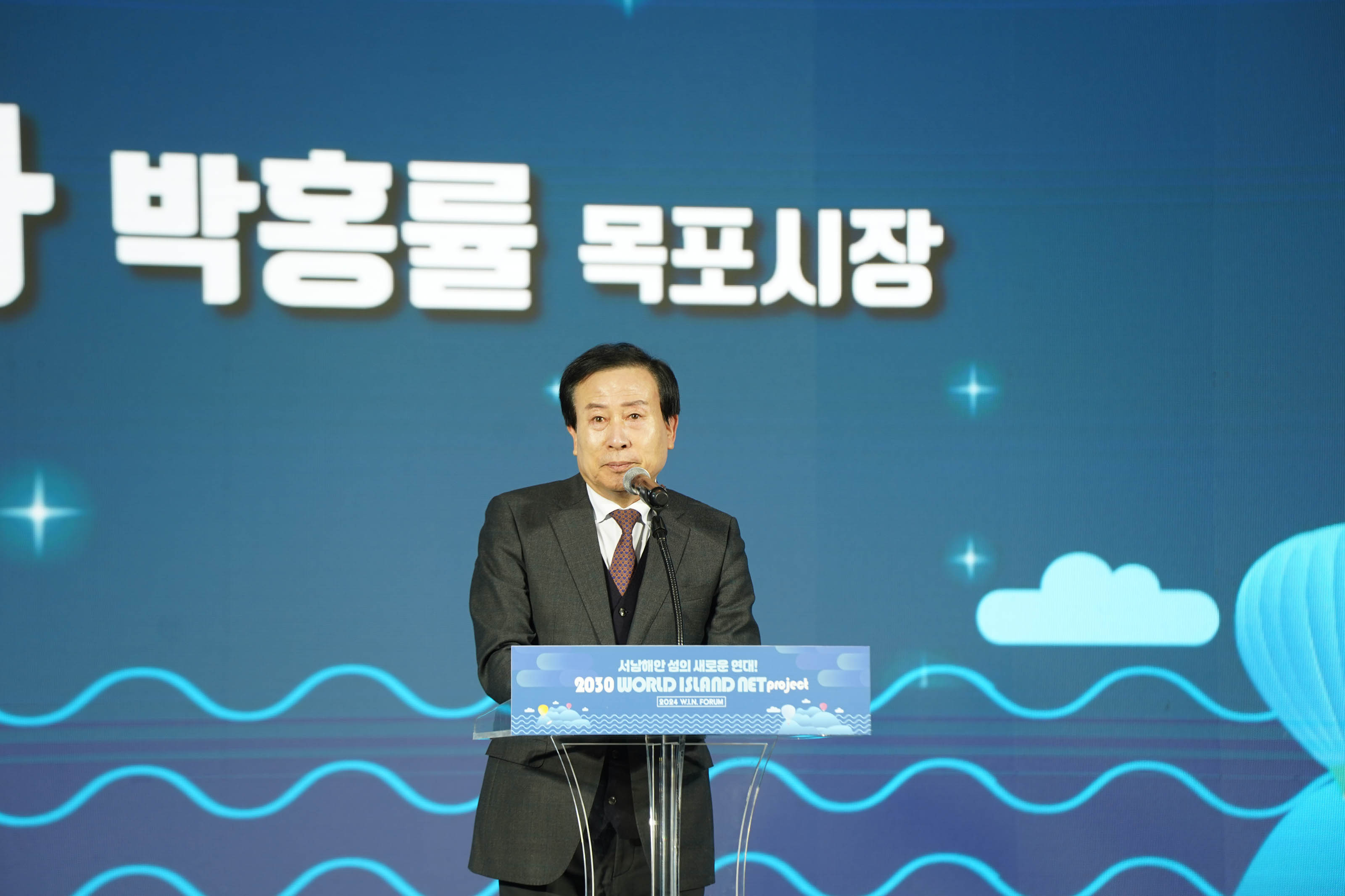 ‘박홍률 목포시장’이라는 문구가 적힌 파란색 배경 스크린 앞 단상에서 정장을 입은 박홍률 시장이 마이크를 들고 세계 W.I.N 포럼에서 연설하고 있는 모습. 단