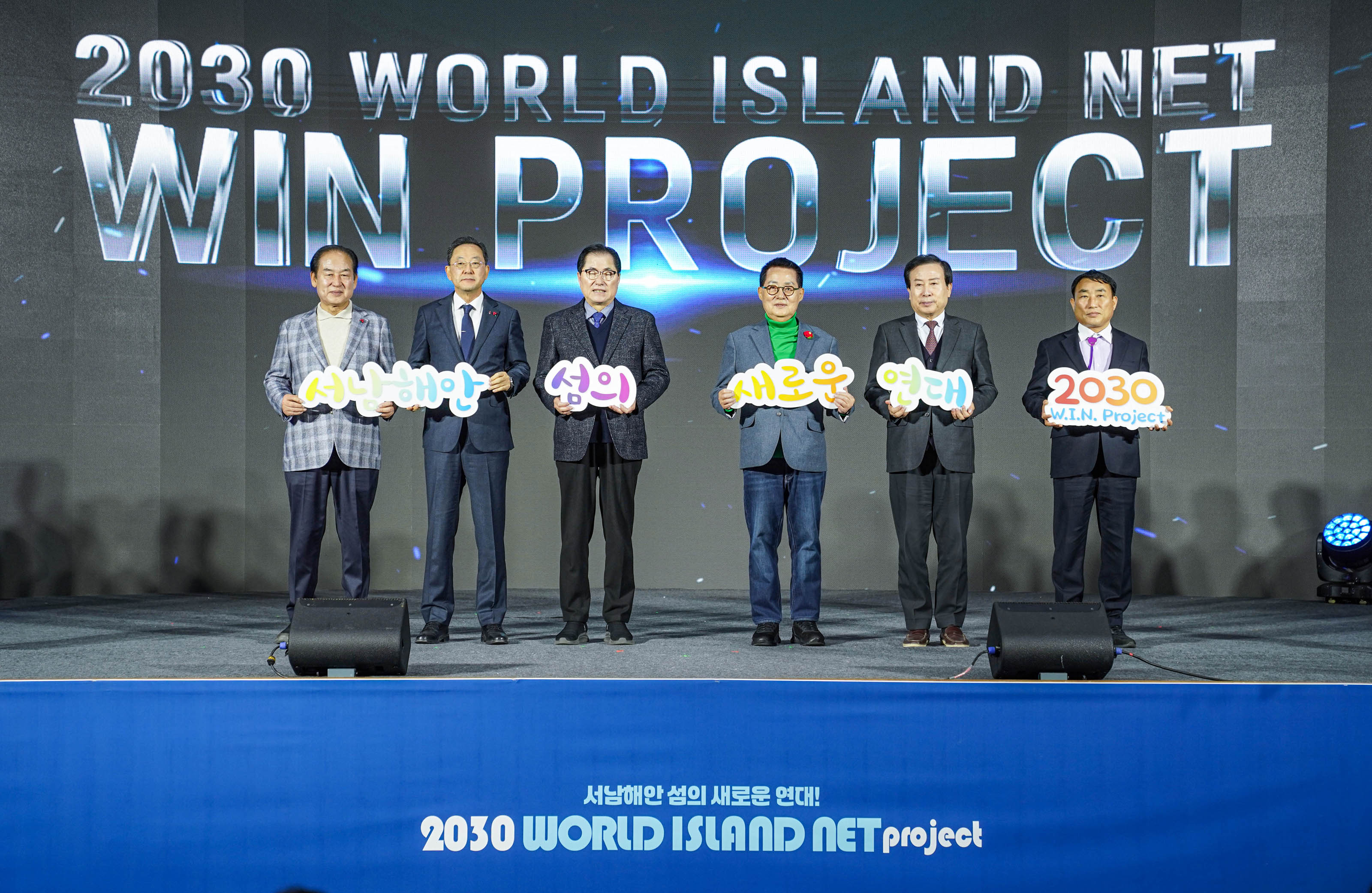 ‘2030 WORLD ISLAND NET WIN PROJECT’라는 대형 배경 화면 앞 무대에서 여섯 명의 관계자가 ‘서남해안’, ‘섬의’, ‘새로운’, ‘연대’, ‘2030 W.I.N. Project’라는 팻말을 각각 들고 일렬로 서 있다. 무대 앞 하단에는 ‘서남해안 섬의 새로운 연대! 2030 WORLD ISLAND NET project’라는 문구가 적혀 있다. 참여자들은 정장 차림으로 포즈를 취하고 있다.