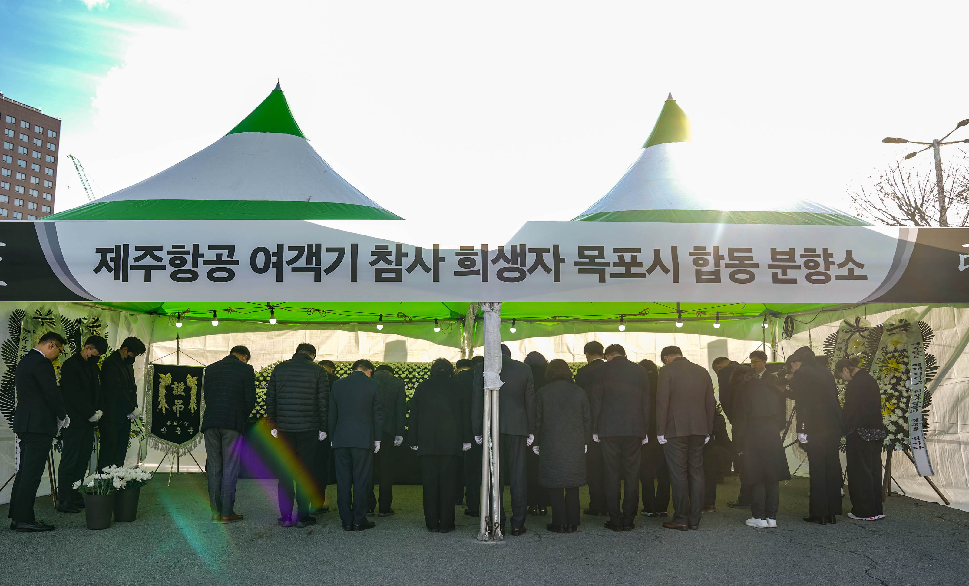 '제주항공 여객기 참사 희생자 목포시 합동 분향소'라는 문구가 적힌 천막 아래, 검은 정장을 입은 20여명의 조문객들이 고개를 숙여 희생자들을 추모하고 있다. 내부에는 국화꽃과 근조기를 비롯한 분향소가 마련되어 있다.