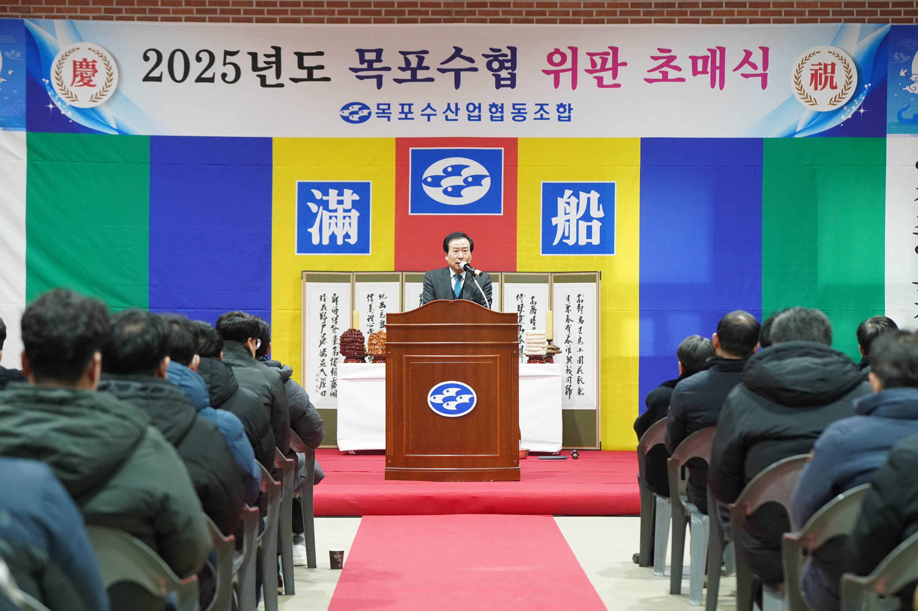 ‘2025년도 목포수협 위판 초매식 목포수산업협동조합’이라는 문구가 적힌 현수막 앞 단상에서 박홍률 목포 시장이 마이크를 잡고 연설하고 있다. 단상 뒤에는 수협 로고와 함께 ‘滿船’이라는 한자가 적힌 문구가 보이며, 단상 앞에는 수십명의 청중들이 경청하는 모습이 담겨 있다.