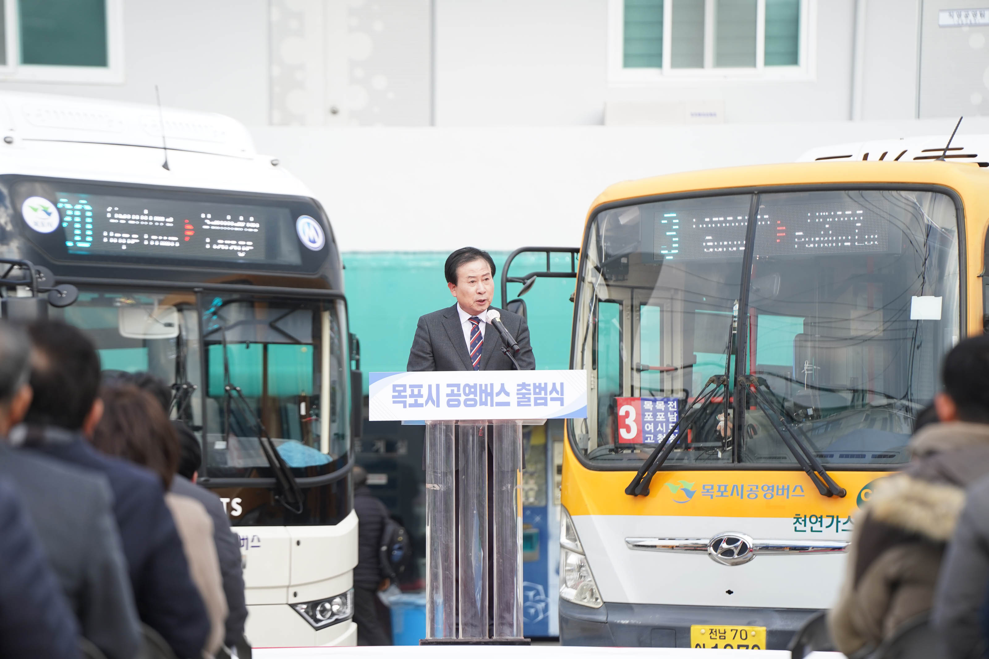목포시 공영버스 출범식에서 박홍률 목포시장이 연설을 하고 있다. 단상 앞쪽에는 '목포시 공영버스 출범식' 이라는 문구가 적혀 있고 참석자들은 단상을 바라보며 의자에 앉아있다. 버스 두 대가 행사 현장에서 전시되어 있다.