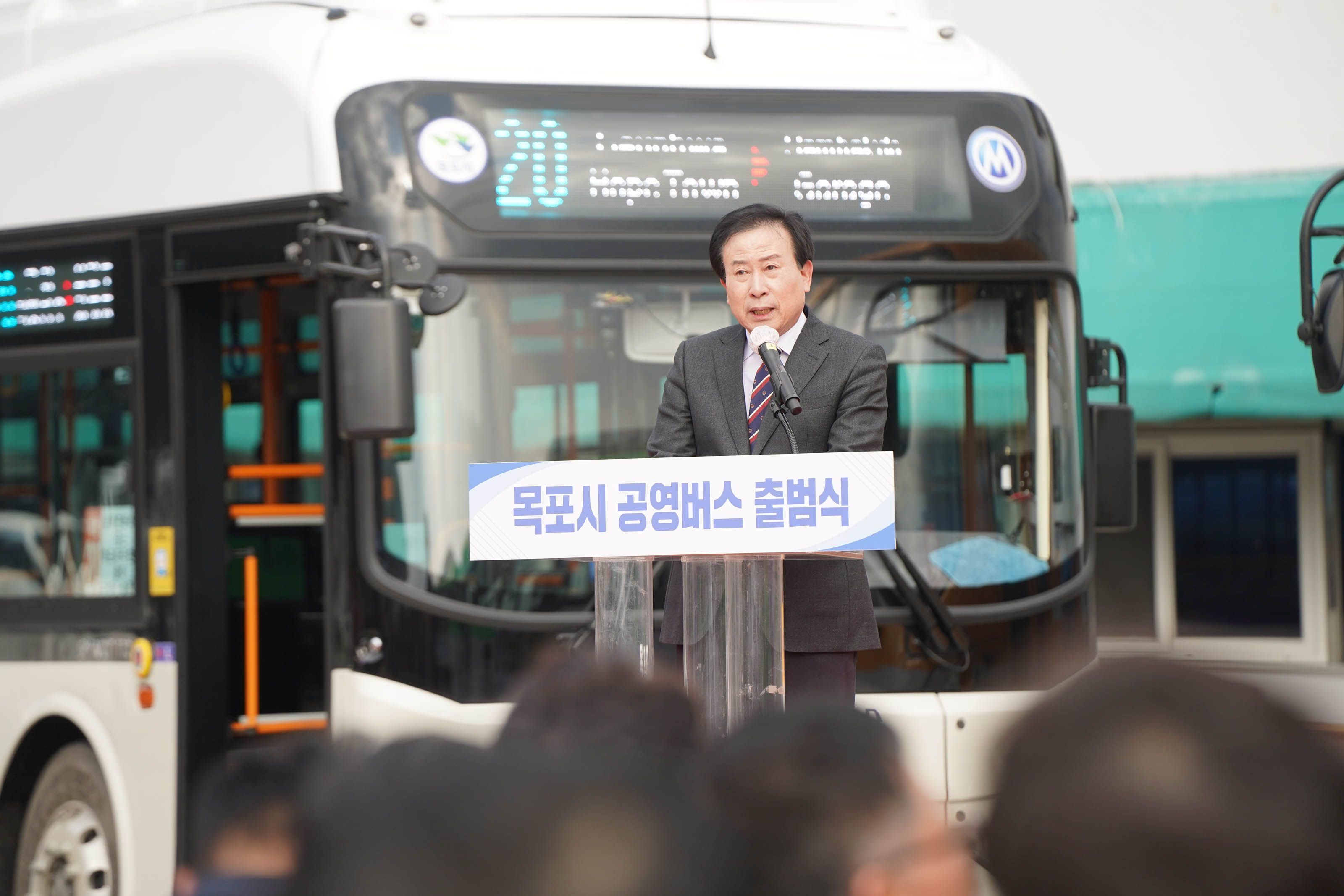 목포시 공영버스 출범식에서 박홍률 목포시장이 연설을 하고 있다. 단상 앞쪽에는 '목포시 공영버스 출범식' 이라는 문구가 적혀 있고 뒷편에는 20번 버스 한 대가 보인다.