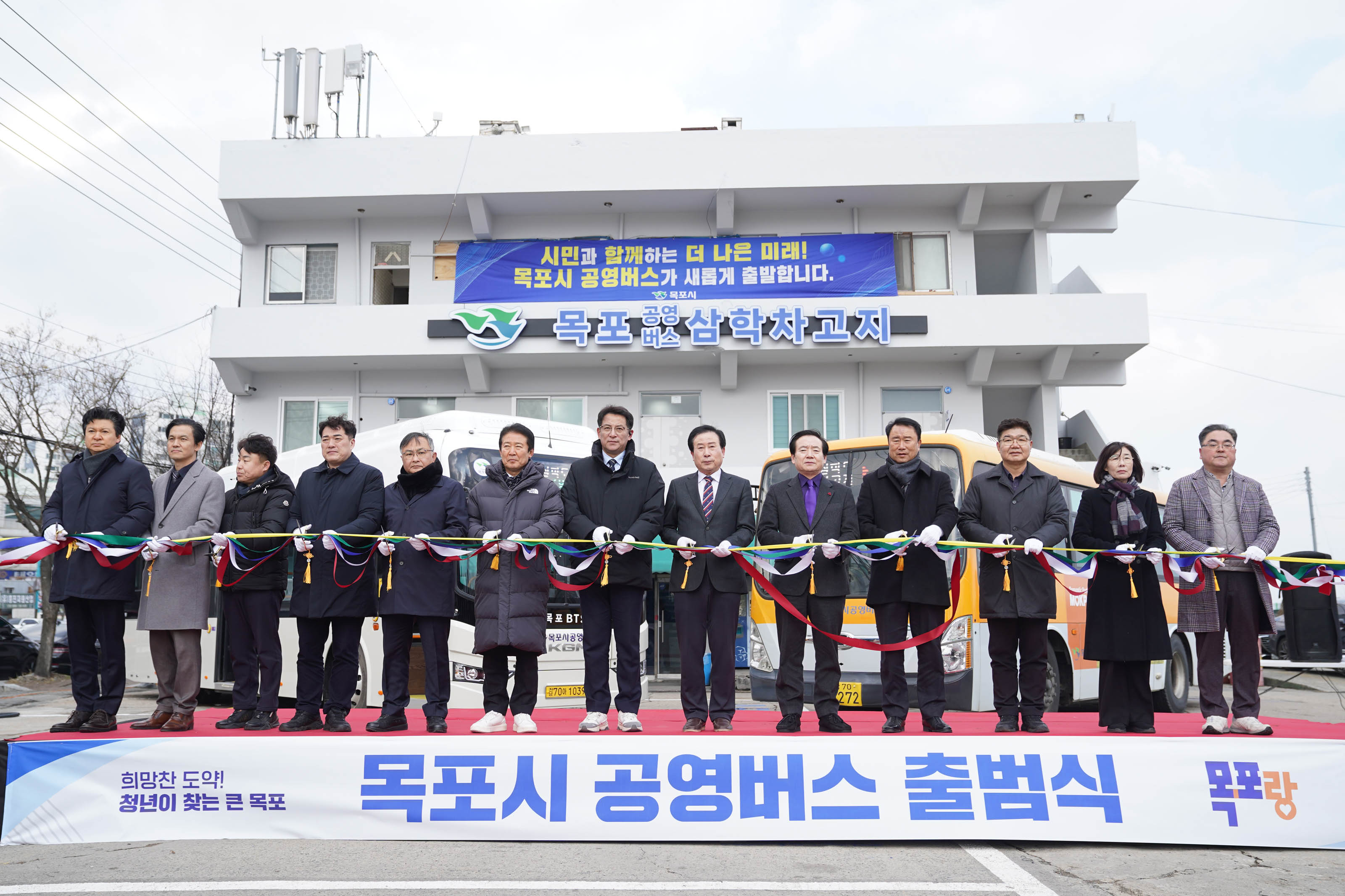 '시민과 함께하는 더 나은 미래! 목포시 공영버스가 새롭게 출발합니다. 목포시'라는 문구가 적힌 현수막이 걸려있는 목포 공영버스 삼학차고지 건물 앞에서 박홍률 목포시장을 포함한 13명의 주요 인사들이 리본을 자르고 있다.