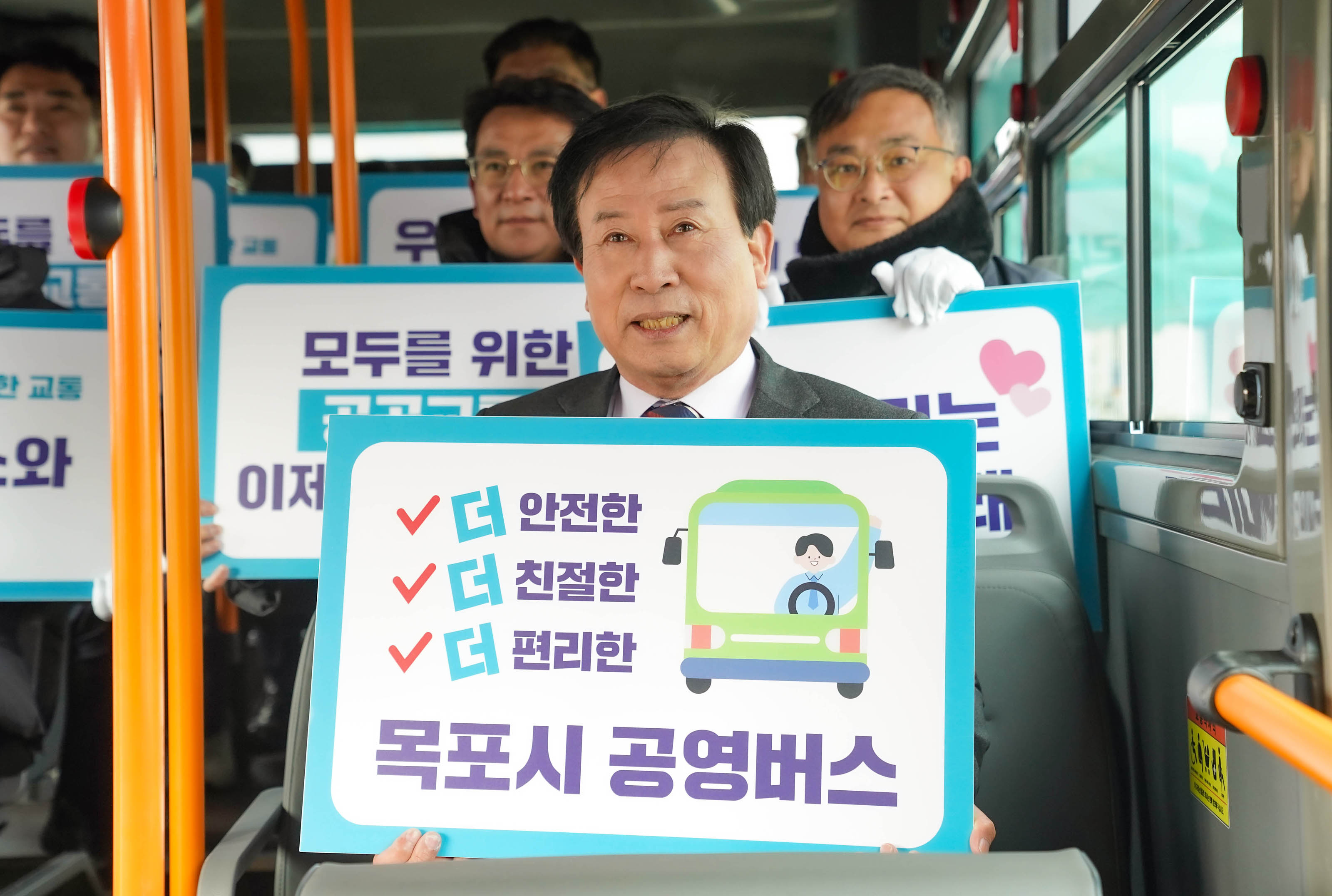 목포시 공영버스 출범식에서 박홍률 목포시장이 버스 안에서 '더 안전한, 더 친절한, 더 편리한 목포시 공영버스'라는 문구가 적힌 팻말을 들고 웃으며 사진을 찍고 있다. 뒷편에는 주요인사들이 팻말을 들고 웃고 있는 모습이 보인다.