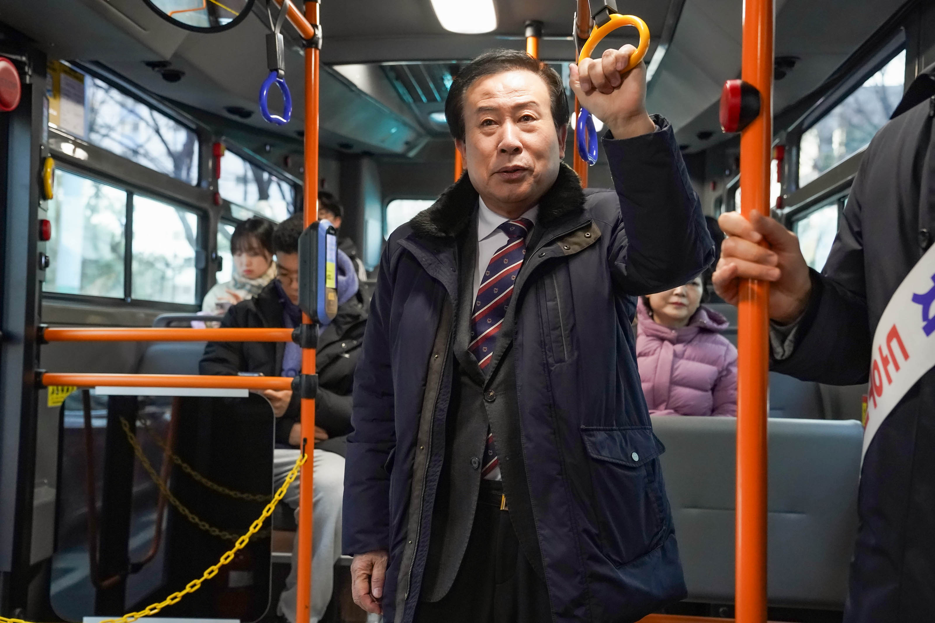 박홍률 목포시장이 공영버스 안에서 손잡이를 잡고 서 있는 모습. 버스 내에는 몇 명의 승객이 앉아 있다.