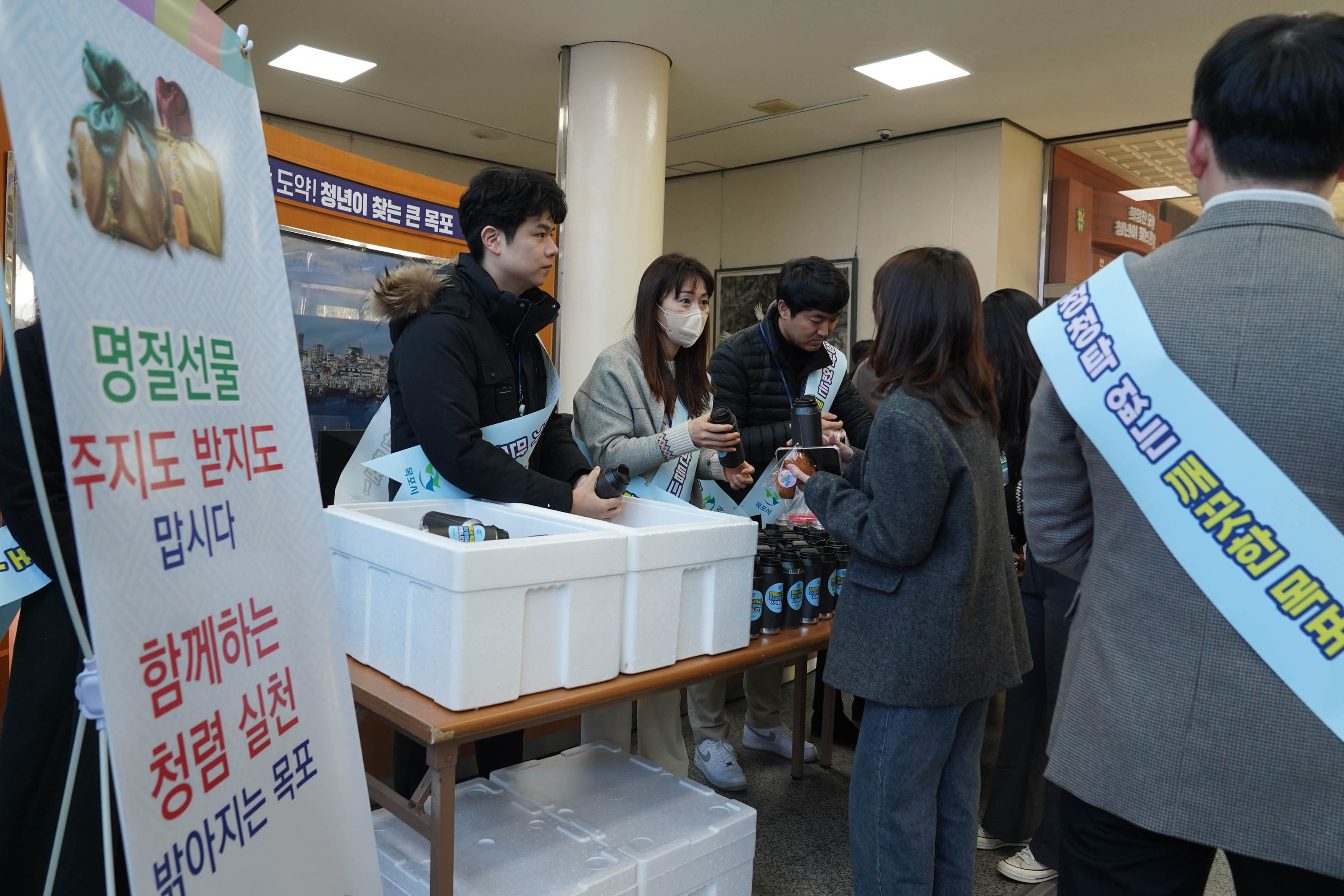 '명절선물 주지도 받지도 맙시다 함께하는 청렴 실천 밝아지는 목포' 라는 문구가 적힌 배너가 왼편에 위치하고 있고 직원들이 진열된 텀블러를 나누어주고 있다. 직