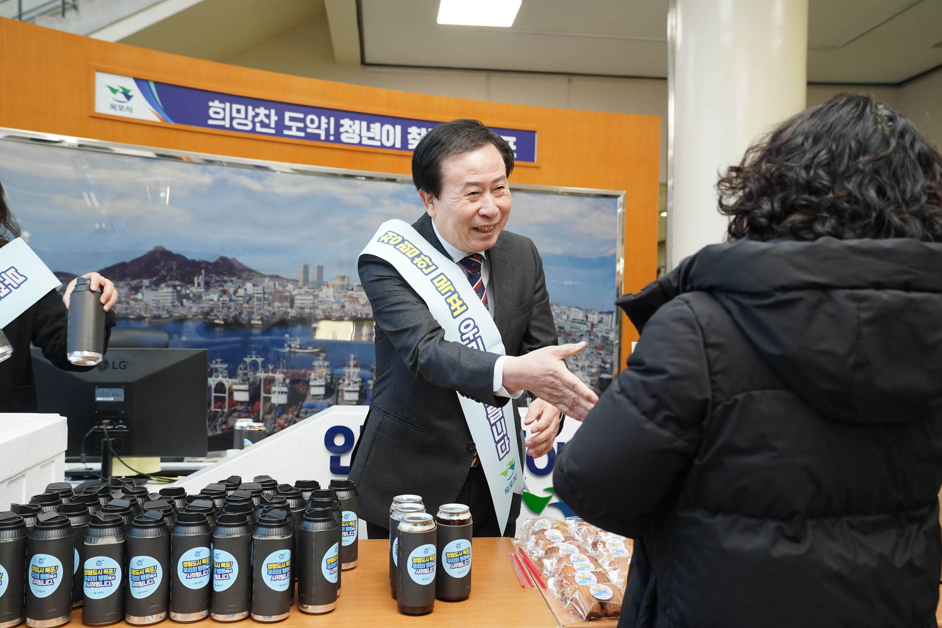 박홍률 목포시장이 청렴 캠페인 물품을 나누며 시민에게 악수를 청하는 모습이다. 테이블에는 수십개의 검은색 텀블러가 놓여 있고, 시장은 캠페인 리본을 착용하고 시민과 친근하게 소통하고 있다.