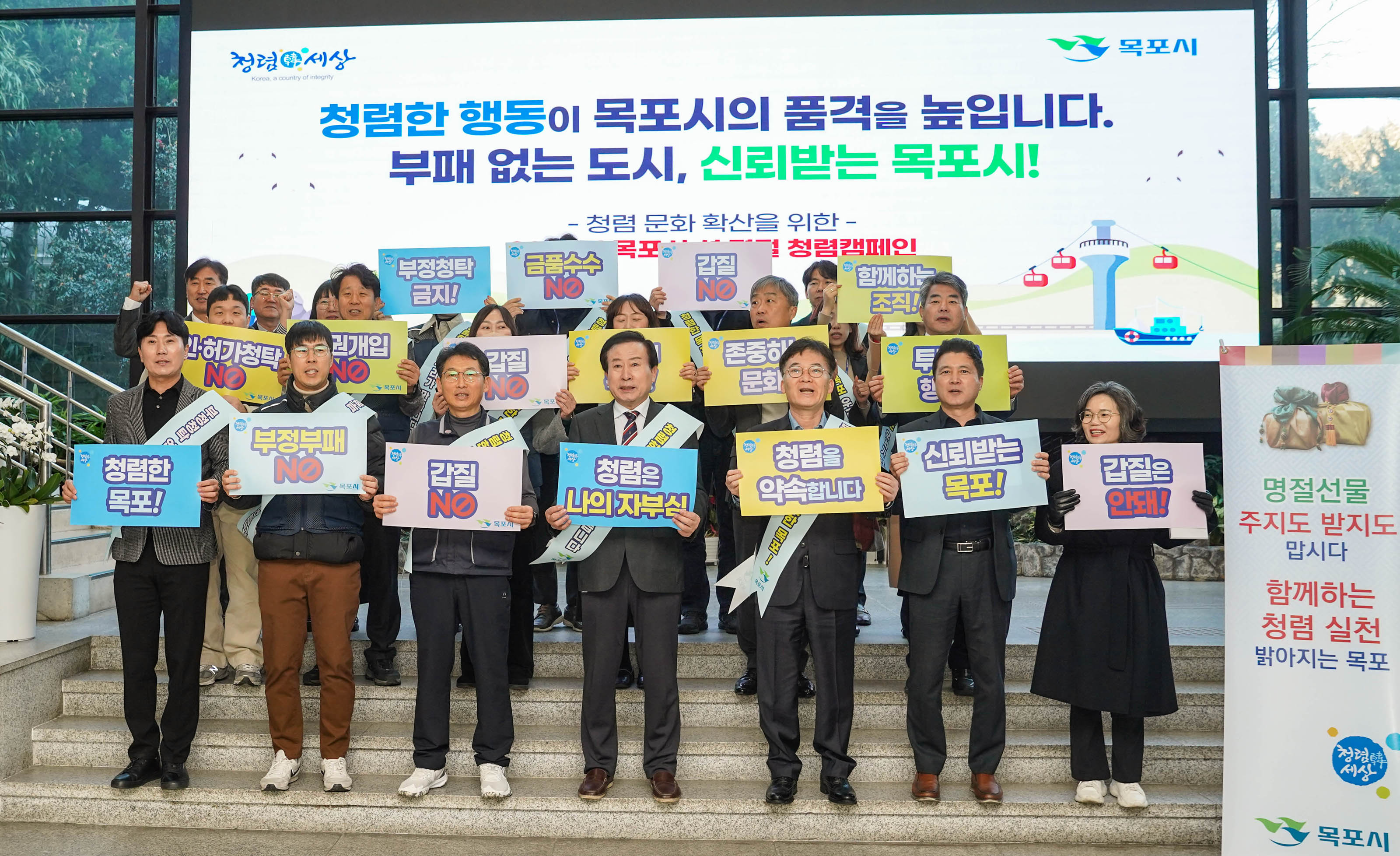 '청렴한 행동이 목포시의 품격을 높입니다. 부패 없는 도시, 신뢰받는 목포시!' 라는 문구가 적힌 배경앞에서 가운데 박홍률 목포시장을 중심으로 20여명의 관계자들이 패킷을 들고 포즈를 취하고 있다. 패킷에는 '청렴한 목포!' , '부정부패 NO','갑질 NO', '청렴은 나의 자부심', '청렴을 약속합니다.' , '신뢰받는 목포!', '갑질은 안돼!' 등의 문구가 적혀 있다.