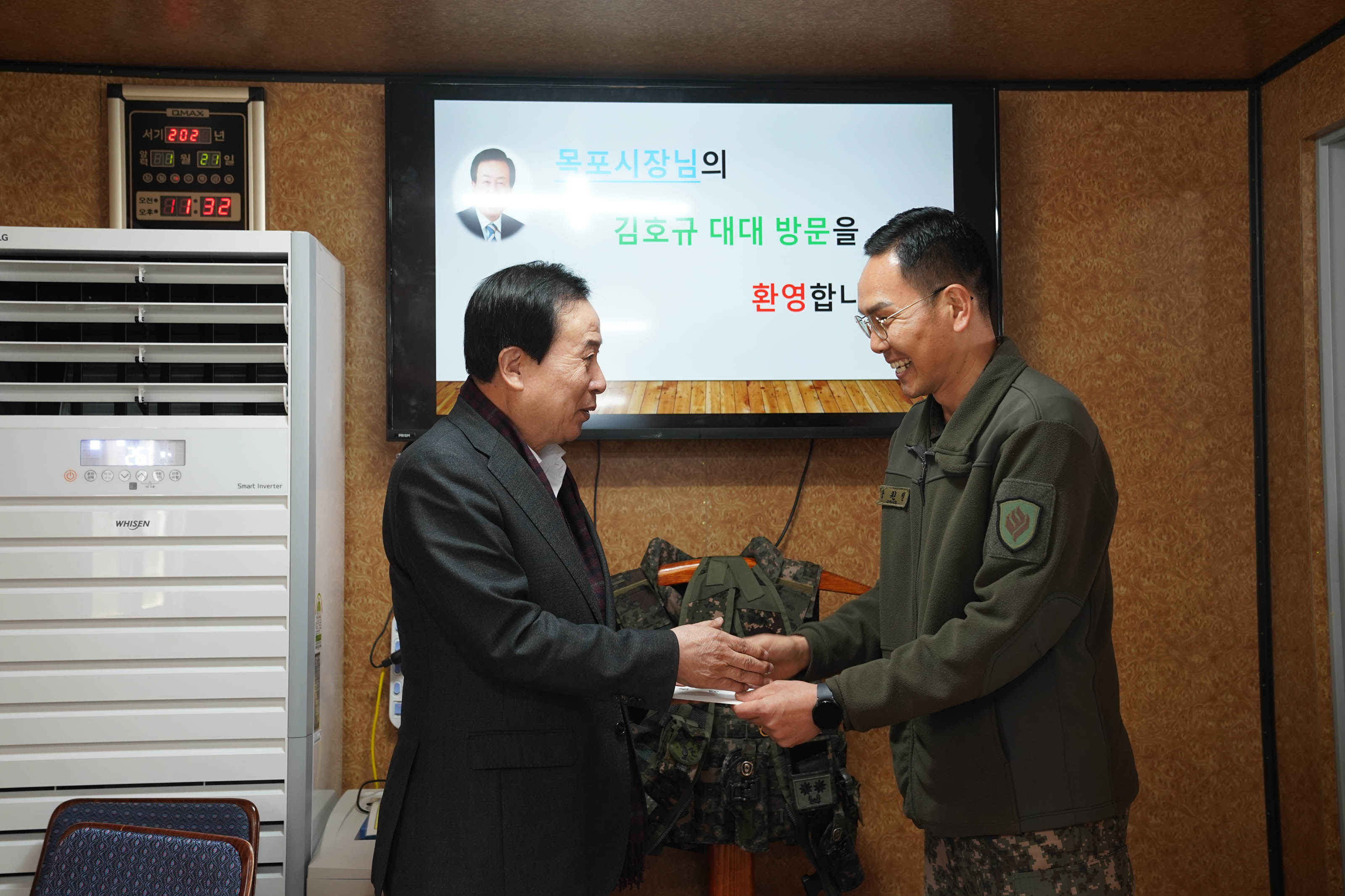 박홍률 목포시장이 군대 부대를 방문하여 군인과 악수를 나누며 환하게 웃고 있다. 화면에는 '목포시장님의 김호규 대대 방문을 환영합니다'라는 환영 메시지가 표시되어 있다. 군인은 군복을 착용하고 있으며, 방 안에는 군용 장비가 배경에 있다.
