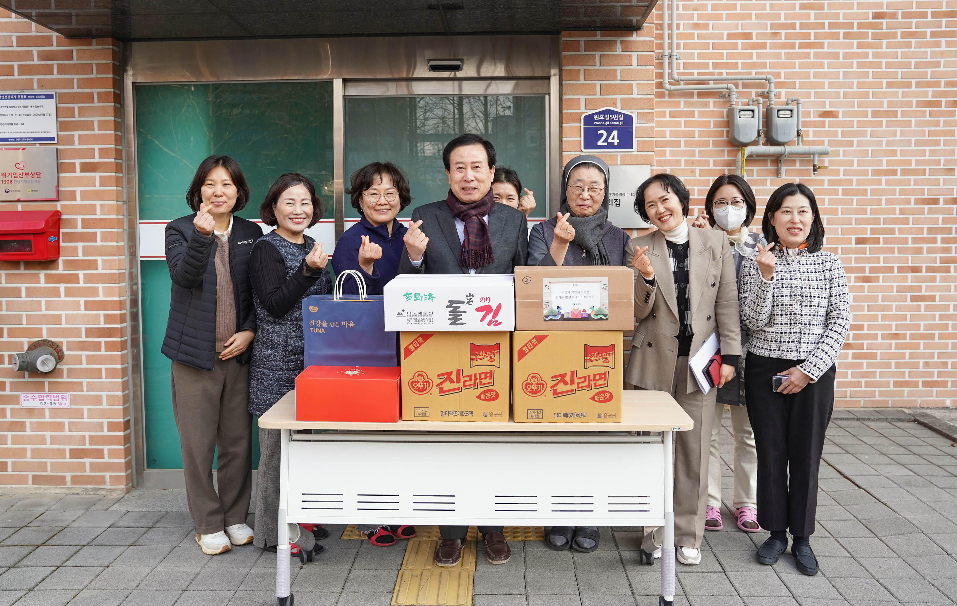 박홍률 목포시장이 8명의 여성 관계자들과 함께 손하트를 하며 포즈를 취하는 모습이다. 기부 물품으로는 박스와 라면 상자들이 보이며 테이블에 박스채로 올려져있다. 직원들은 모두 웃으며 손하트를 하고 있고, 시장은 가운데에서 직원들과 함께 미소를 지으며 서 있다.
