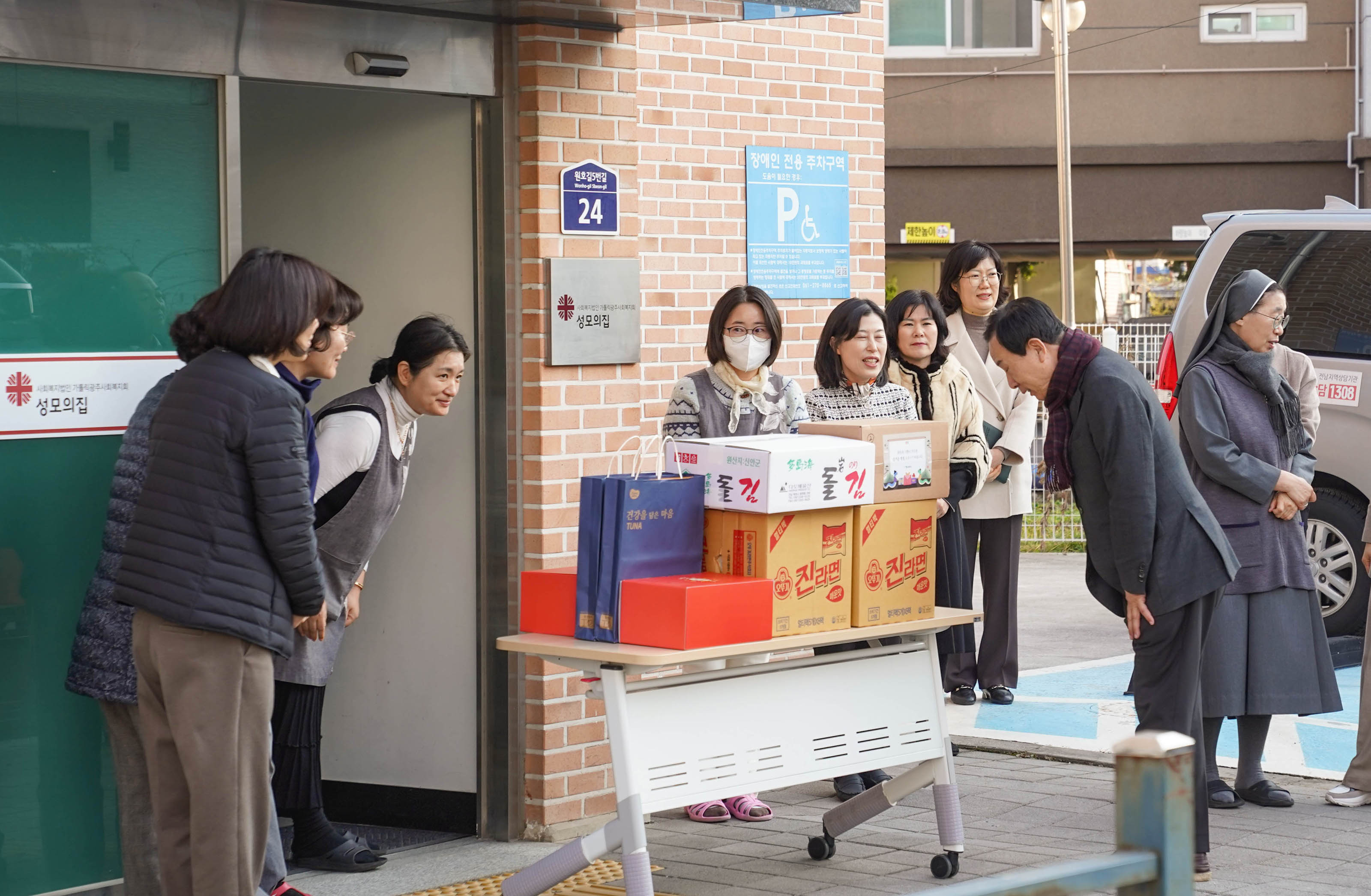 박홍률 목포시장이 기부 물품을 전달하고, 직원들과 인사를 나누는 모습이다. 기부 물품으로는 박스와 라면 상자들이 보이며, 직원들은 기부를 받은 후 미소를 지으며 인사를 나누고 있다. 시장은 손을 숙여 인사를 하고 있으며, 9명의 직원들은 손을 모아 겸손히 대답하는 모습이다. 