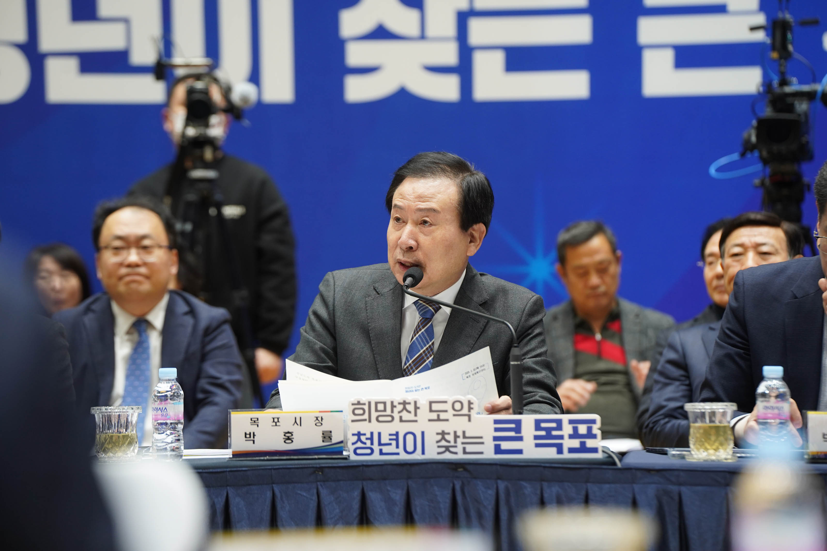 정장 차림의 박홍률 목포시장이 마이크 앞에서 발언하고 있는 모습. 앞에는 이름표가 있으며, 이름표 상단에는 '목포시장 박홍률'이라고 적혀 있고, 아래쪽에는 '희망찬 도약! 청년이 찾는 큰 목포'라는 문구가 큼직하게 표시되어 있음. 배경에는 '청년이 찾는 큰 목포'라는 대형 문구가 파란색 배경에 새겨져 있고, 뒤쪽에는 여러 명의 관계자들이 앉아 있으며 일부는 노트북을 사용하고 있음.