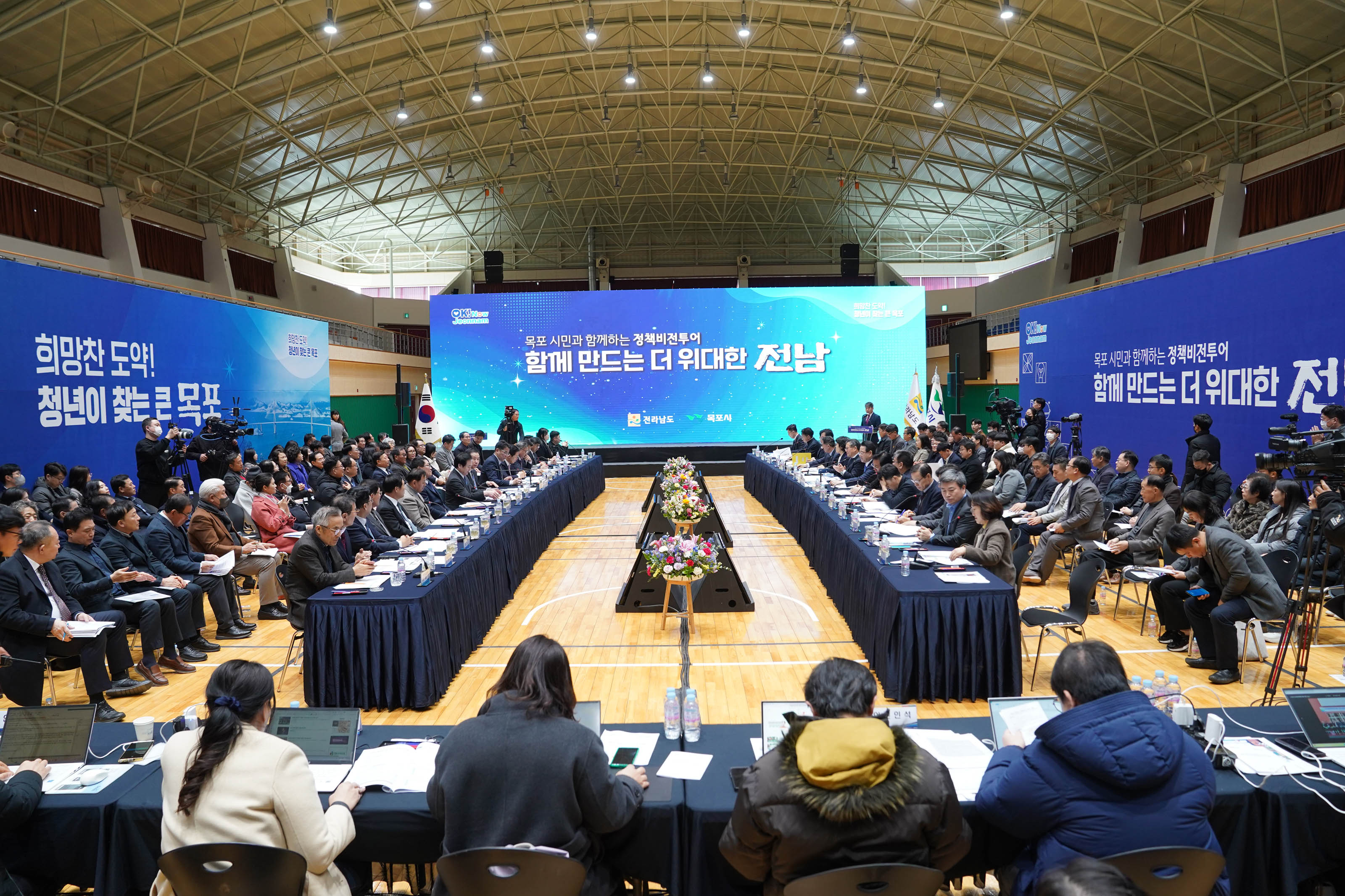 실내 체육관 형태의 장소에서 열린 대형 회의 현장. 중앙에는 꽃으로 장식된 긴 테이블이 있고, 양쪽에 회의 참가자들이 길게 둘러앉아 있음. 왼쪽 배경에는 파란색 벽면에 '희망찬 도약! 청년이 찾는 큰 목포'라는 문구가 적혀 있고, 오른쪽 배경에는 '목포 시민과 함께하는 정책비전투어 함께 만드는 더 위대한 전남'이라는 대형 LED 전광판이 설치되어 있음. 중앙에는 'OK! Now Jeonnam'이라는 텍스트도 보임. 관계자들과 참석자들이 진지하게 회의에 임하고 있는 모습.