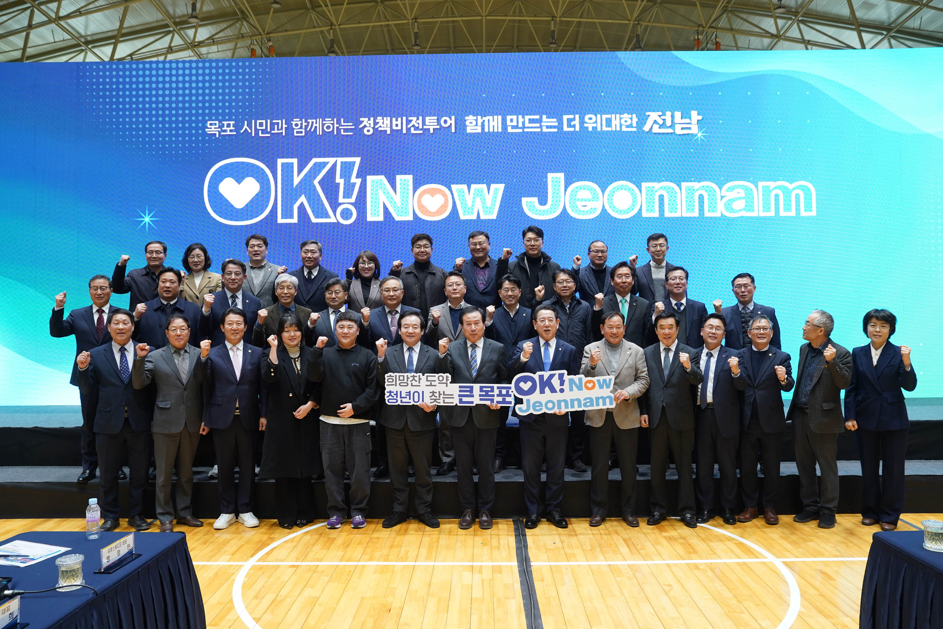 정장을 입은 참석자들이 단체로 무대 위에 모여 기념사진을 촬영하는 장면. 중앙에는 'OK! Now Jeonnam'이라는 대형 LED 스크린이 배경으로 있으며, 그 위에 '목포 시민과 함께하는 정책비전투어 함께 만드는 더 위대한 전남'이라는 문구가 큼직하게 표시되어 있음. 앞줄 중앙에는 박홍률 시장이 있으며, '희망찬 도약! 청년이 찾는 큰 목포'와 'OK! Now Jeonnam'이라고 적힌 팻말을 들고 기념촬영 중. 모든 인원이 밝은 표정으로 손을 들어 파이팅 자세를 취하고 있음.