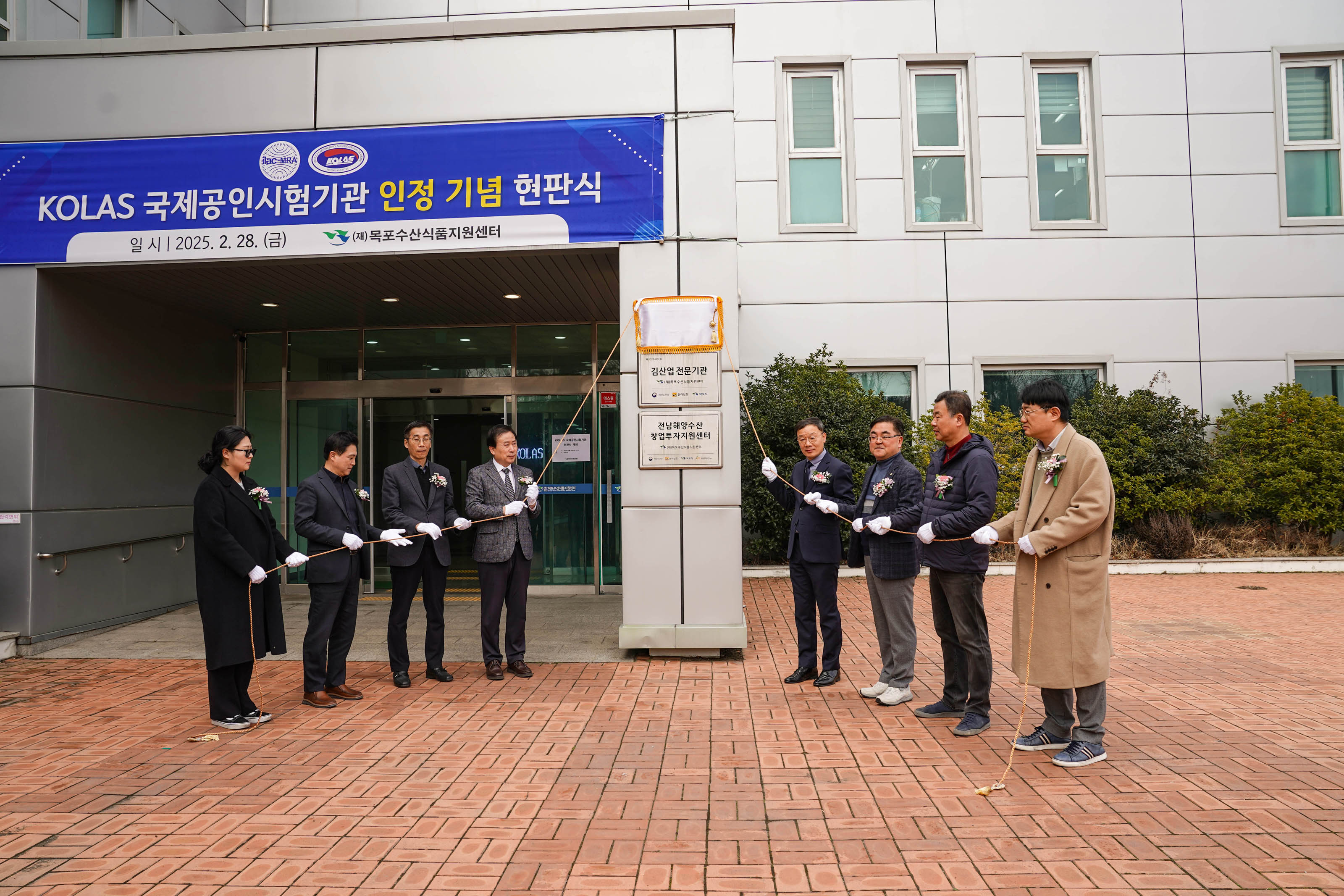 건물 앞에서 박홍률 목포시장을 비롯한 정장 차림의 관계자들이 하얀 장갑을 끼고 리본을 잡고 서 있는 현판식 장면. 건물 입구 상단 현수막에는 다음과 같은 문구가 있음
'KOLAS 국제공인시험기관 인정 기념 현판식
일 시 | 2025. 2. 28. (금)
(재)목포수산식품지원센터'
