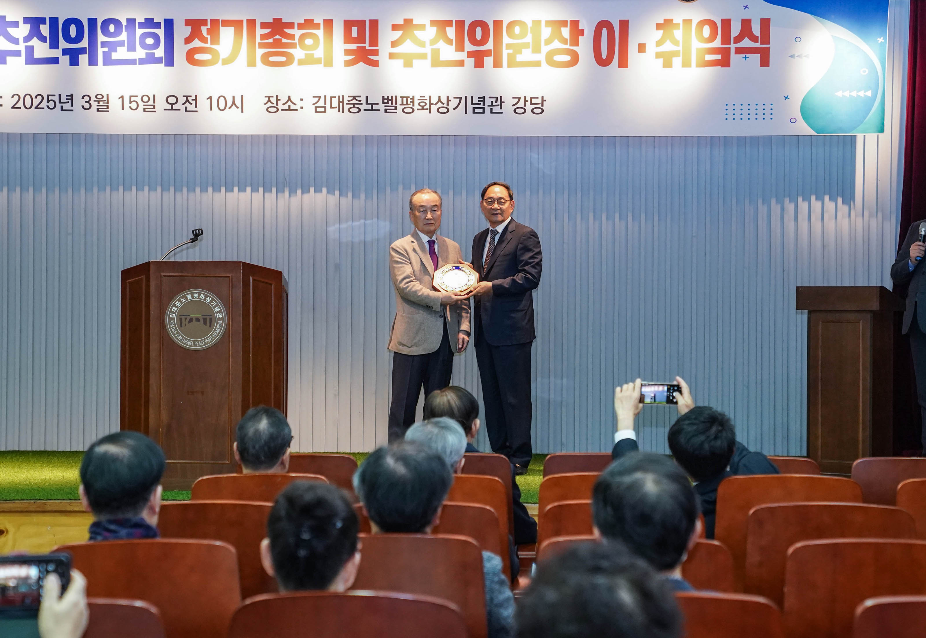 목포신안통합추진위원회 정기총회 및 추진위원장 이·취임식에서 관계자들이 접시를 들고 사진을 찍는 모습. 배경에는 '목포신안통합추진위원회 정기총회 및 추진위원장 이·취임식'이라는 배너가 보이고, 그 아래에는 '일시: 2025년 3월 15일 오전 10시'와 '장소: 김대중노벨평화상기념관 강당'이 적혀 있다. 청중들이 자리에 앉아 이 장면을 보고 있다.