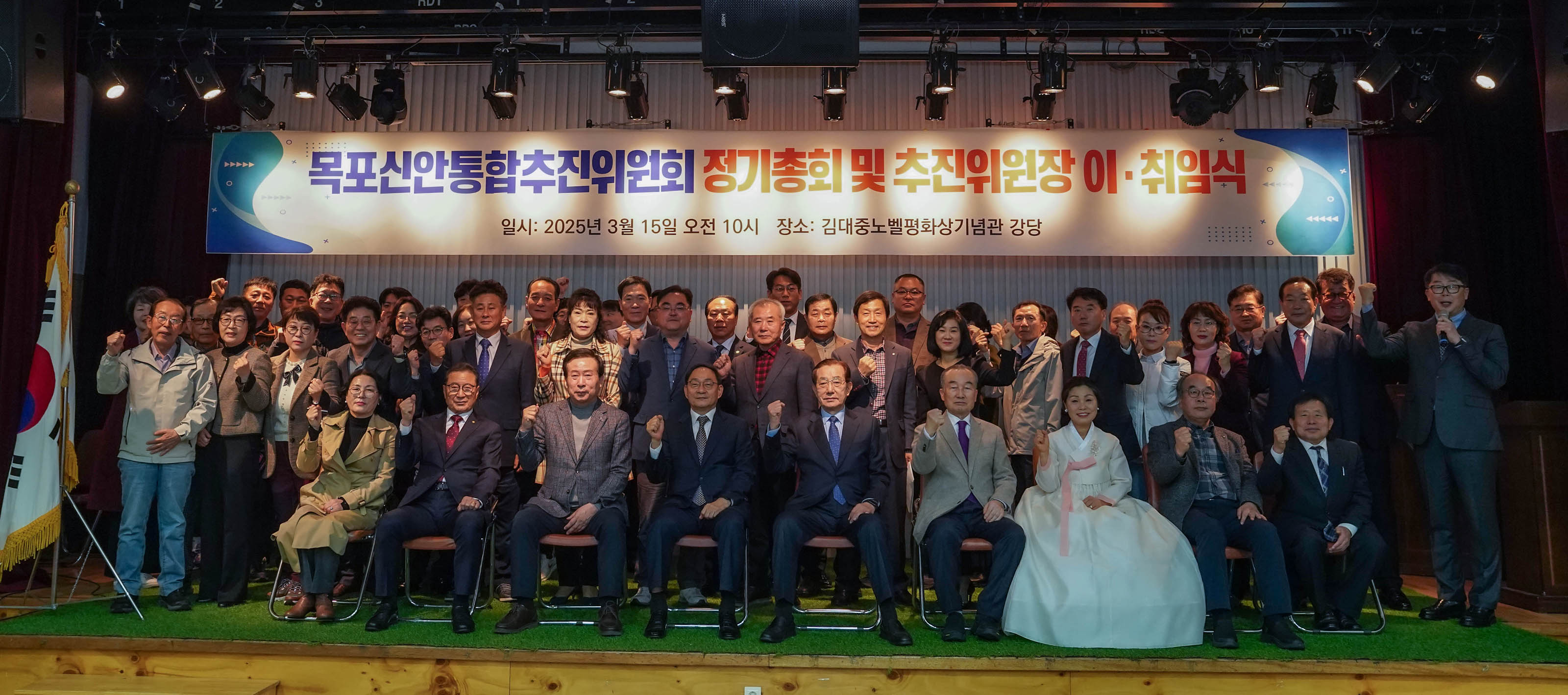 목포신안통합추진위원회 정기총회 및 추진위원장 이·취임식에 참석한 사람들의 단체 사진. 배경 배너에는 '목포신안통합추진위원회 정기총회 및 추진위원장 이·취임식'이라는 문구가 보이며, 그 아래에는 '일시: 2025년 3월 15일 오전 10시'와 '장소: 김대중노벨평화상기념관 강당'이 적혀 있다. 참석자들은 손을 들어 응원의 포즈를 취하고 있다.