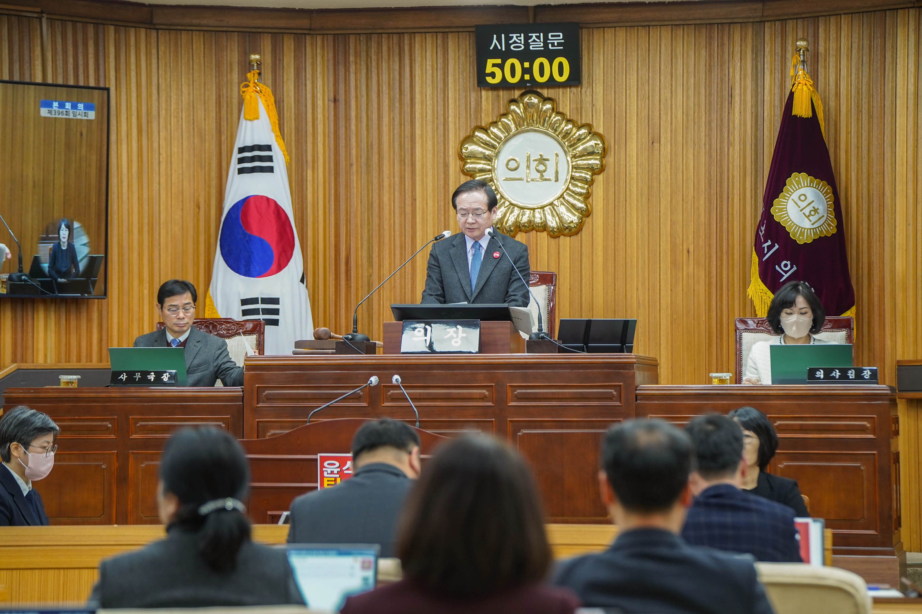 회의실에서 진행 중인 의회 회의. 의장석에 앉은 남성이 마이크 앞에서 발언하고 있으며, 뒤에는 대한민국 국기가 걸려 있고, 상단에는 '시정질문 50:00'이라는 타이머가 보인다. 의장석 옆에는 '의장'이라는 이름이 적힌 이름표가 놓여 있으며, 오른쪽에는 '의사팀장'이 앉아 있다. 앞에는 여러 명이 앉아 있는 모습이 보인다.