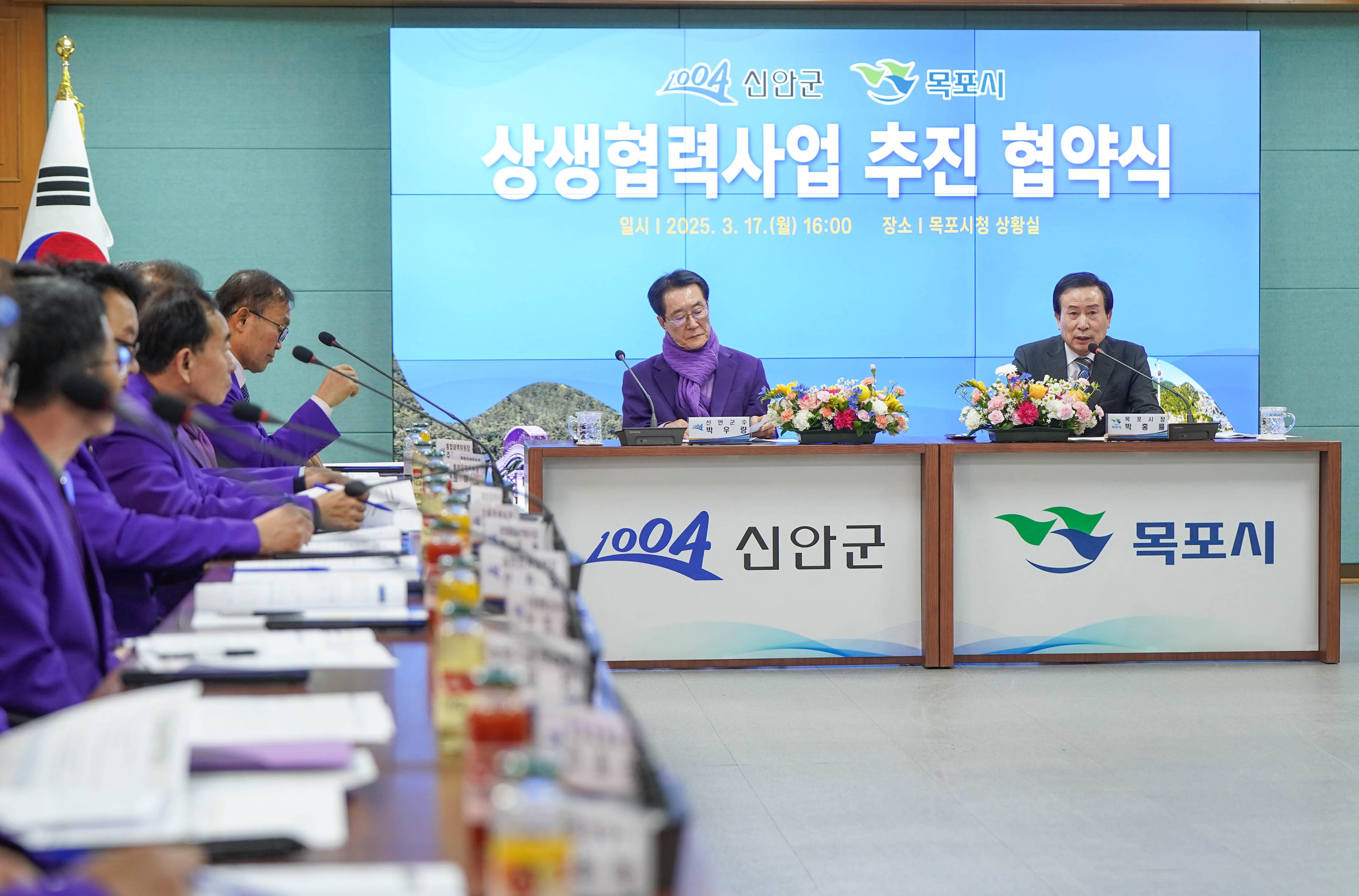 회의 중인 상생협력사업 추진 협약식 장면. 보라색 옷을 입은 관계자들이 앉아 있으며, 회의 테이블 위에는 꽃과 음료들이 배치되어 있다. 화면에는 '신안군 목포시 상생협력사업 추진 협약식 2025. 3. 17. (월) 16:00 장소| 목포시청 상황실'이라는 날짜와 시간이 표시되어 있다.