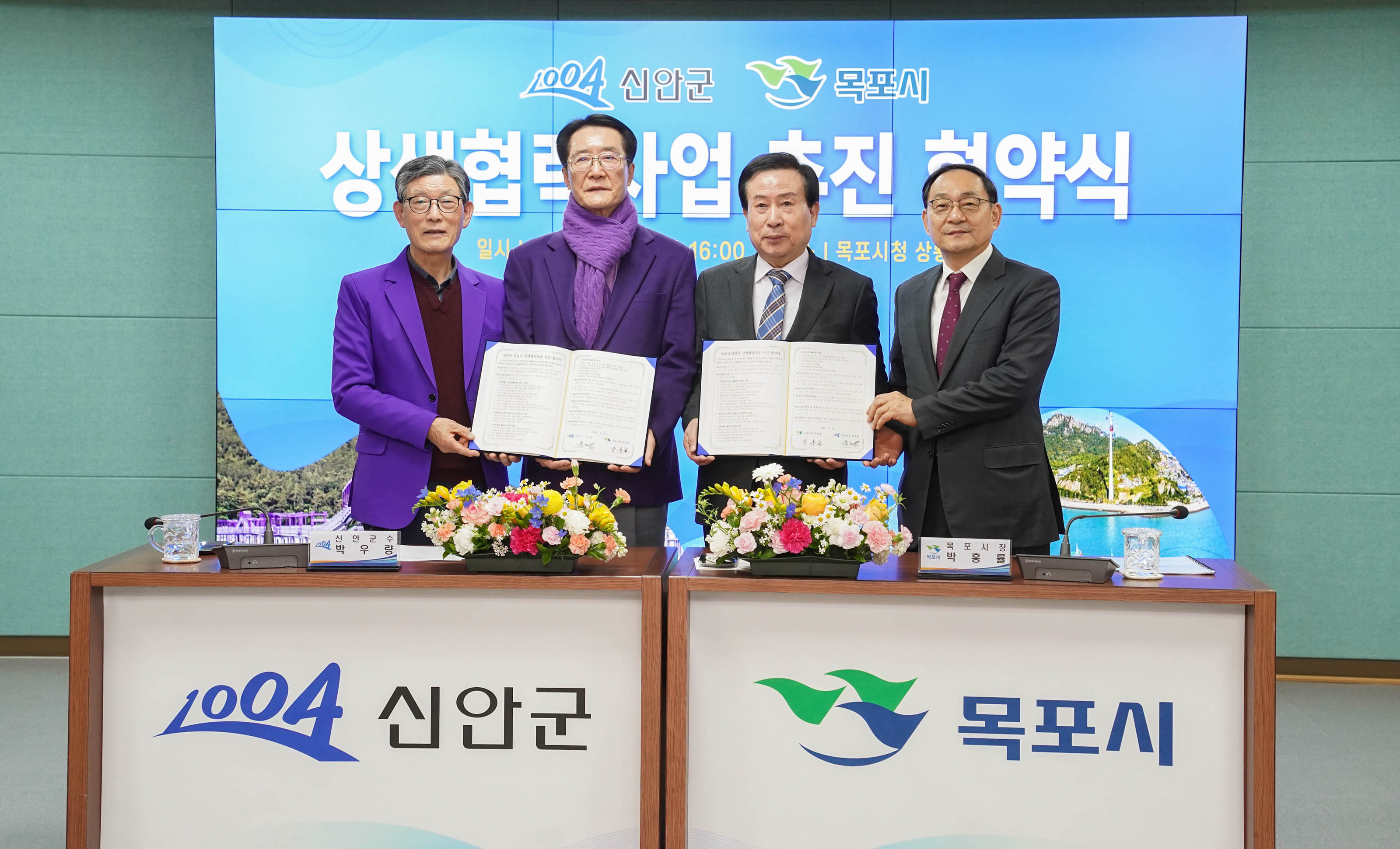 박홍률 목포시장과 세 명의 관계자들이 협약서를 들고 있는 모습. 화면에는 '신안군 목포시 상생협력사업 추진 협약식'이라는 문구가 보이고, 테이블 위에는 꽃이 놓여 있고 테이블에는 1004 신안군 목포시 라는 문구가 적혀있다.