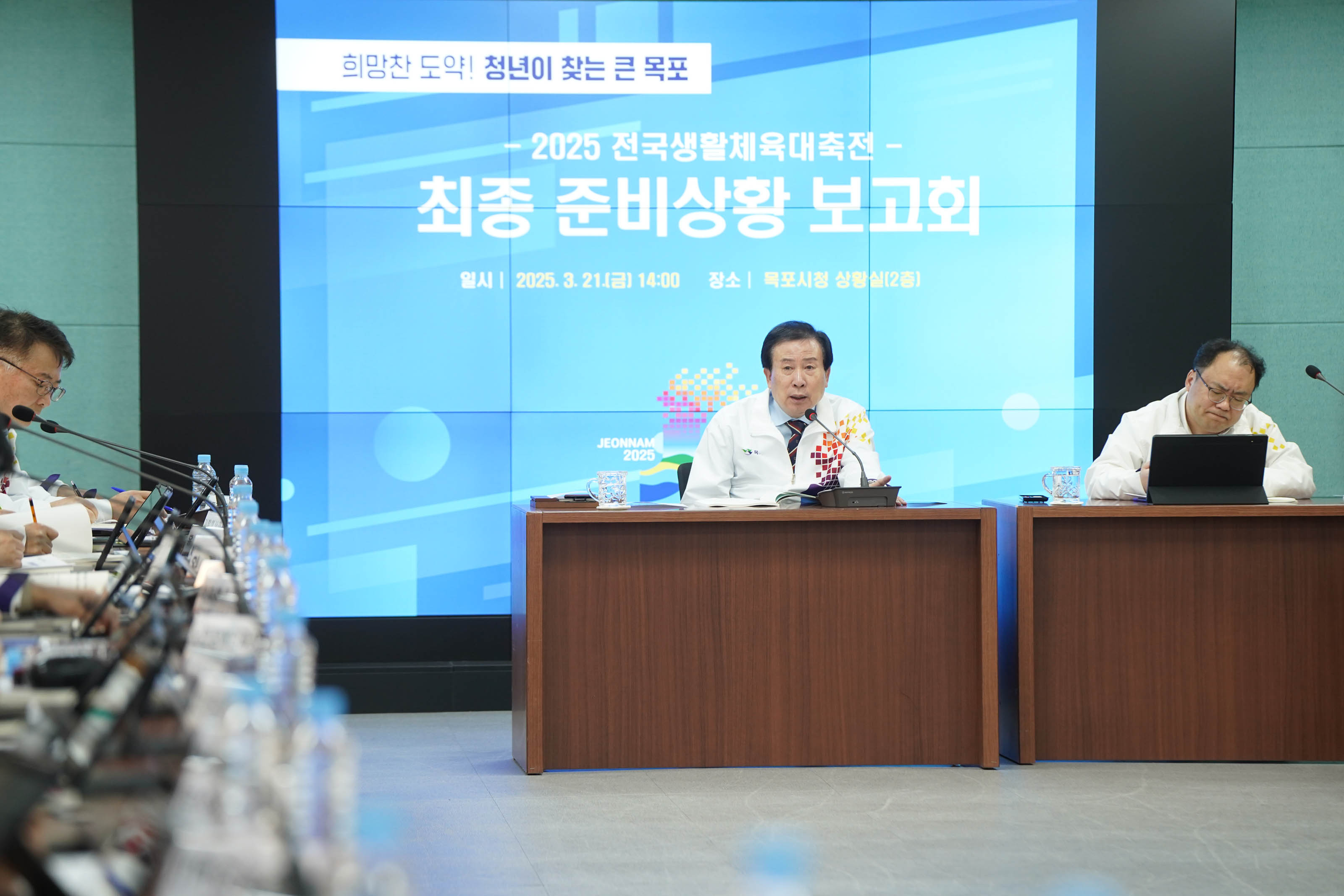 '희망찬 도약! 청년이 찾는 큰 목포 2025 전국생활체육대축전-최종 준비상황 보고회' 라는 문구가 적힌 스크린 앞에서 발표 중인 박홍률 목포시장의 모습. 흰색 자켓을 입고 있으며, 다른 참석자는 주의를 기울이며 발표를 듣고 있다.